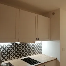 Białe szafki kuchenne z czarnymi uchwytami, mozaika z geometrycznym wzorem nad blatem, płyta indukcyjna i zlewozmywak ze stali nierdzewnej.