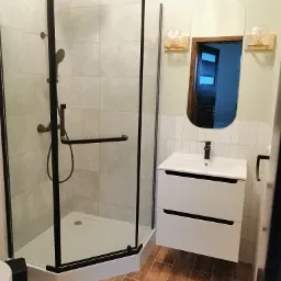 Nowoczesna łazienka z kabiną prysznicową typu walk-in z czarnymi ramami, białą umywalką z szafką, lustrem owalnym z drewnianą ramą i drewnopodobną podłogą z płytek.