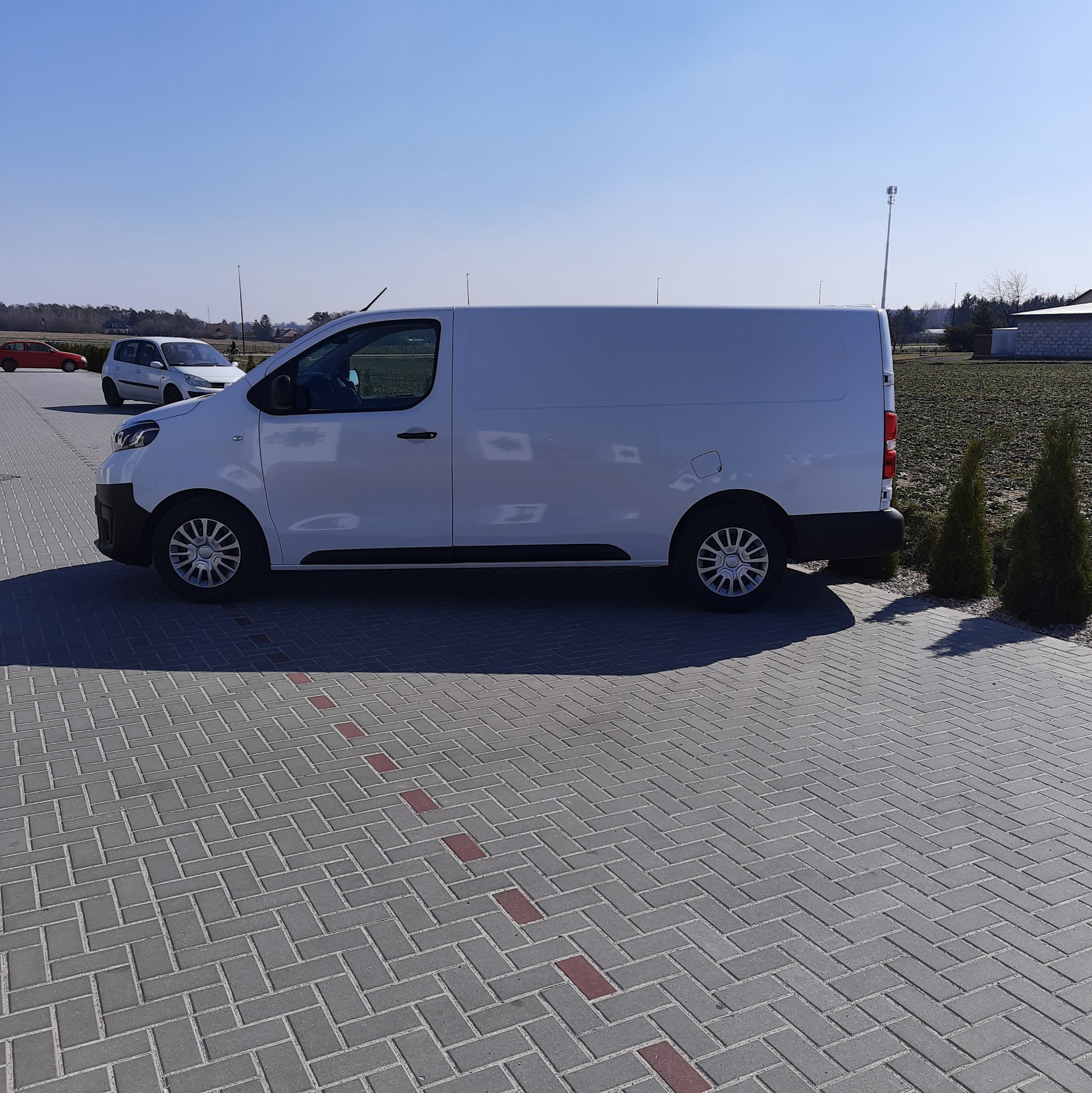 Biały bus dostawczy zaparkowany na kostce brukowej z widocznymi innymi pojazdami w tle, w słoneczny dzień.