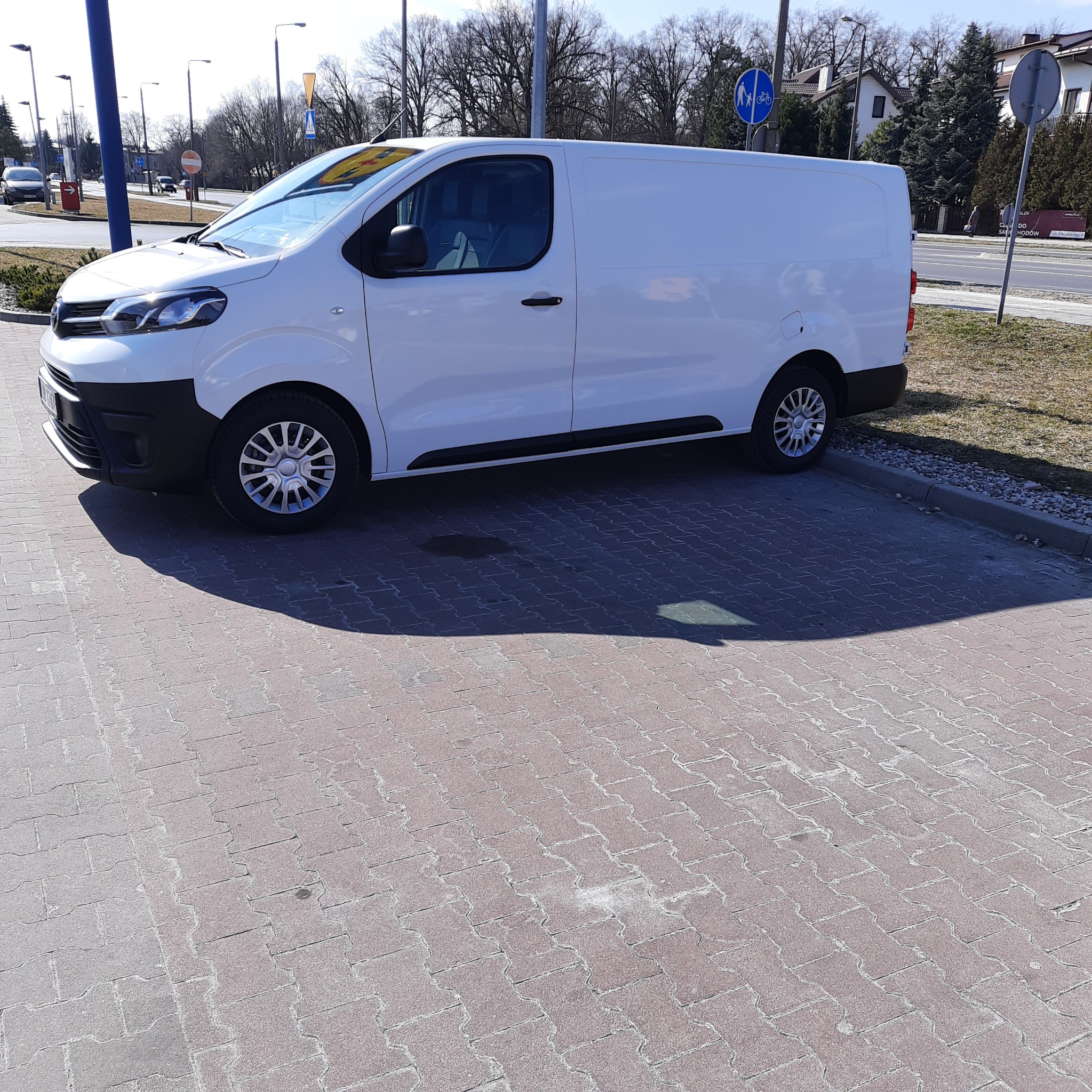 Biały samochód dostawczy zaparkowany na brukowanym parkingu w słoneczny dzień, widoczne znaki drogowe w tle.