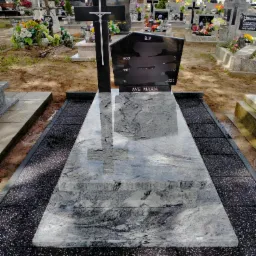 Granitowy nagrobek z krzyżem na cmentarzu, otoczony czarnym grysem. Na płycie napis 'AVE MARIA'. W tle inne nagrobki i zieleń.