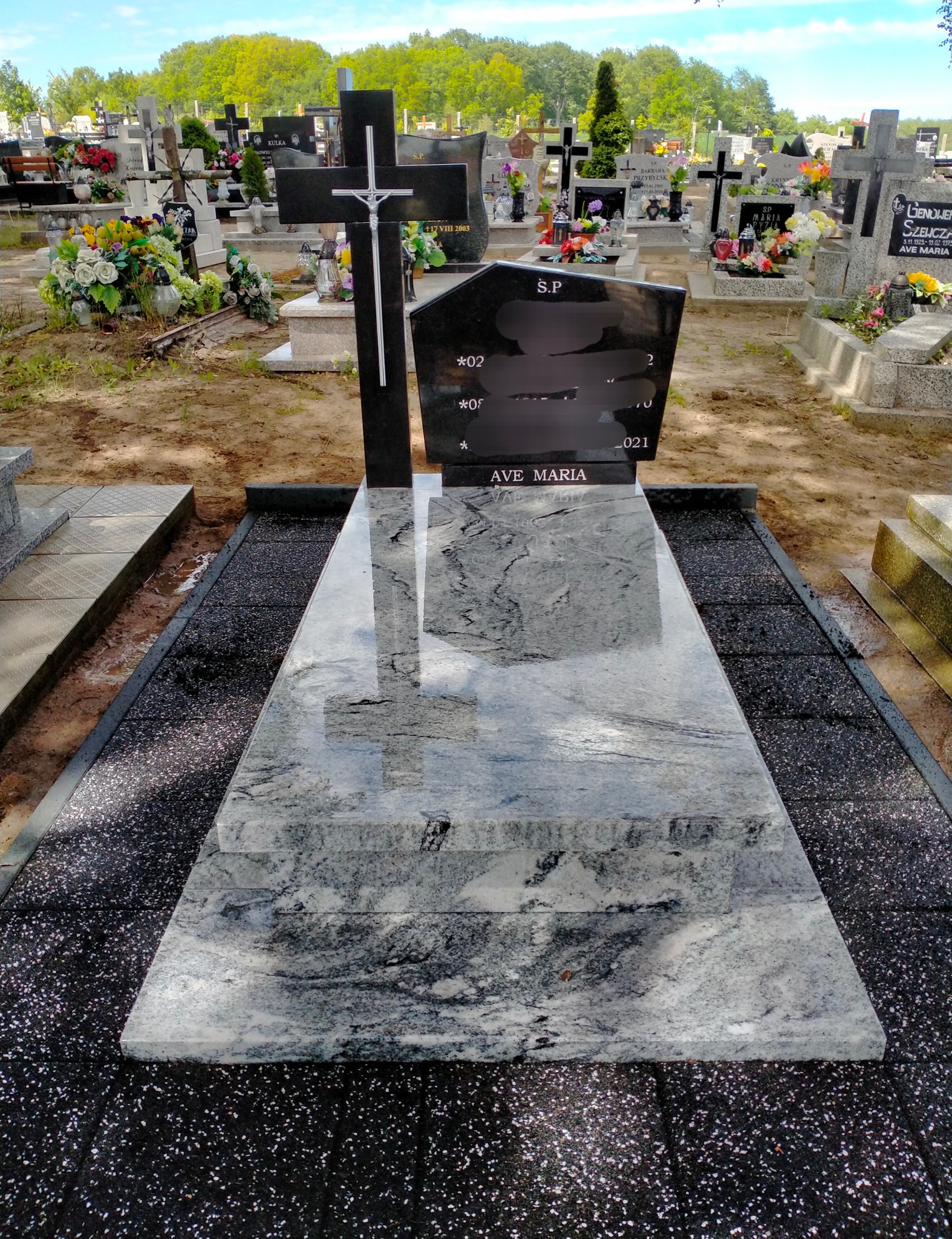 Granitowy nagrobek z krzyżem na cmentarzu, otoczony czarnym grysem. Na płycie napis 'AVE MARIA'. W tle inne nagrobki i zieleń.