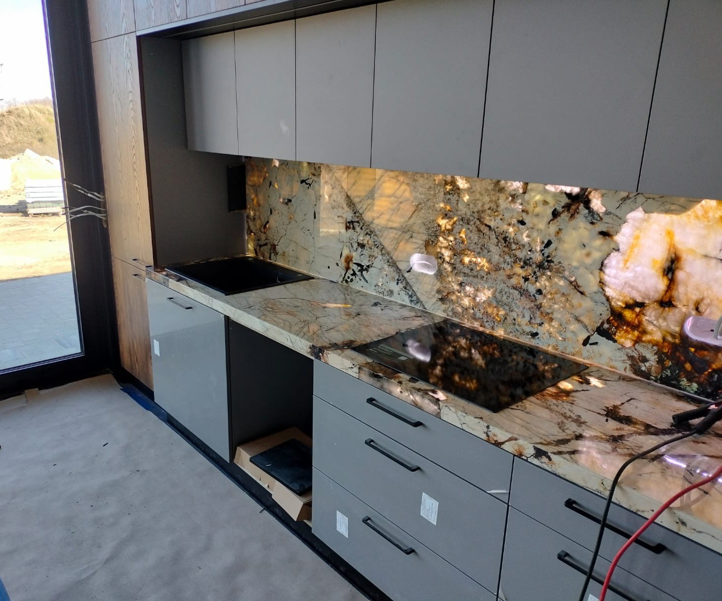 Szara zabudowa kuchenna z blatem i panelem ściennym z kamienia o unikalnym wzorze, z widoczną płytą indukcyjną, w nowoczesnym wnętrzu.