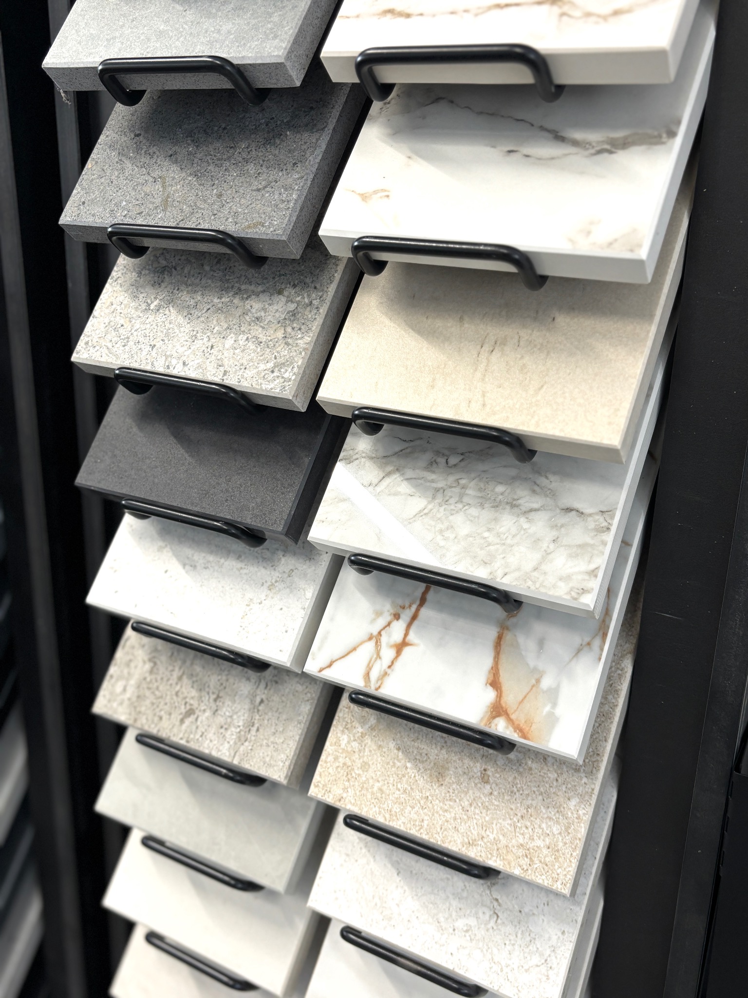 Ekspozycja próbek płytek ceramicznych w różnych kolorach i wzorach, zawieszonych na czarnych metalowych uchwytach, prezentowanych w pionowym układzie.