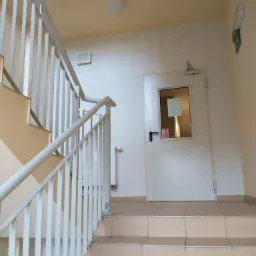 Klatka schodowa z jasnymi płytkami na schodach, białą balustradą i białą ścianą z delikatnym wzorem, prowadząca do białych drzwi z okienkiem.