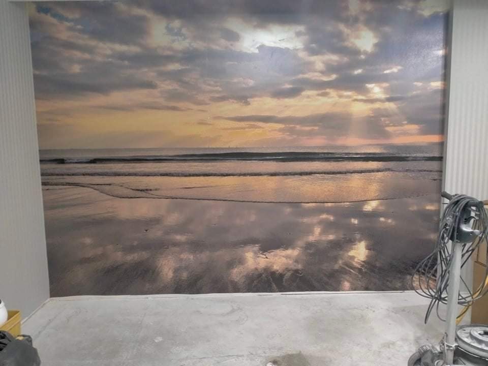 Fototapeta z motywem plaży o zachodzie słońca na ścianie w pomieszczeniu, widoczny fragment podłogi i sprzęt do szlifowania.