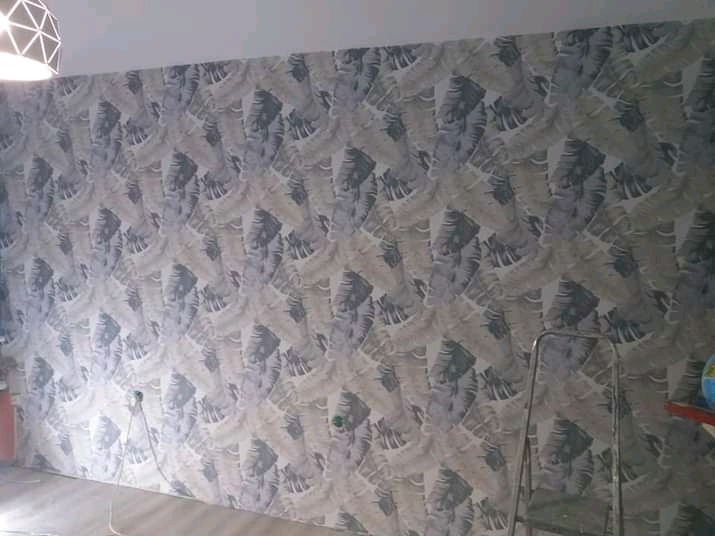 Ściana pokryta tapetą w motywy liści palmowych w odcieniach szarości i bieli, widoczna drabina malarska i fragment lampy sufitowej.