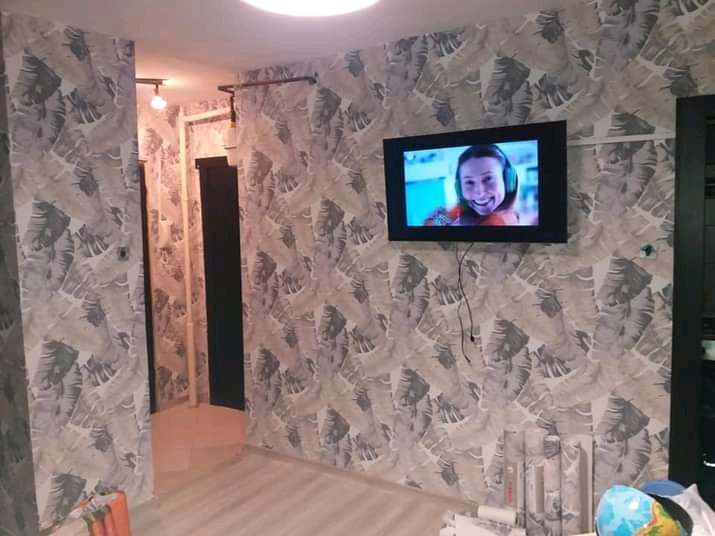 Ściany w pokoju w Kołobrzegu pokryte tapetą w motywy liści, z telewizorem wiszącym na ścianie i rolkami tapety leżącymi na podłodze.