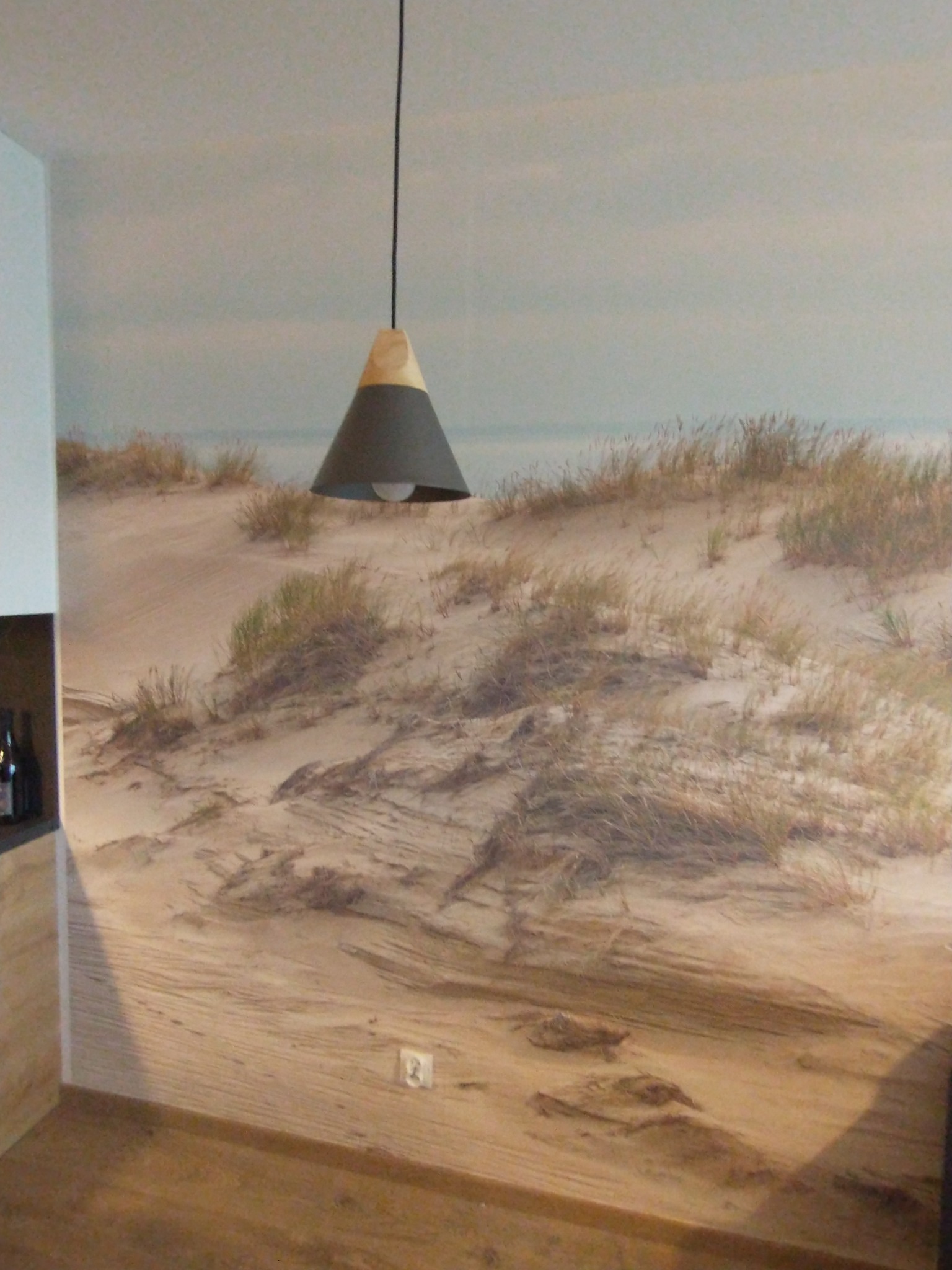 Ściana z fototapetą przedstawiającą wydmy nad morzem, oświetlona stożkową lampą wiszącą z drewnianym elementem, widoczny fragment drewnianej podłogi i gniazdko elektryczne.