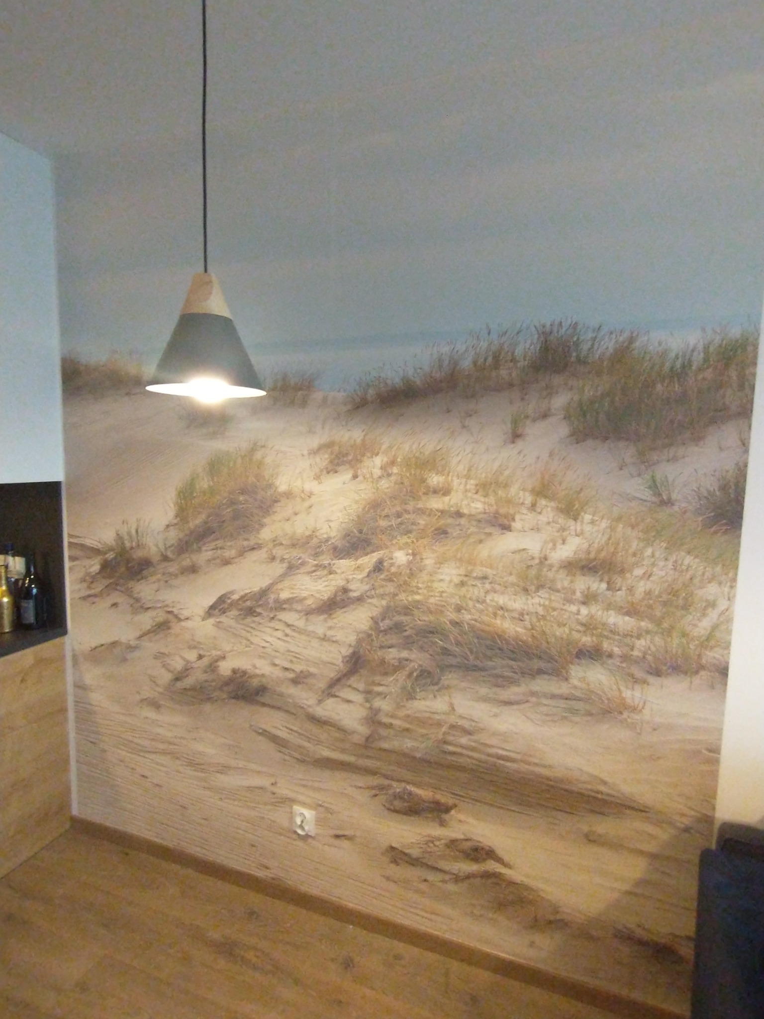 Fototapeta z motywem wydm nadmorskich w oświetlonym wnętrzu, z widoczną lampą wiszącą o stożkowym kloszu.