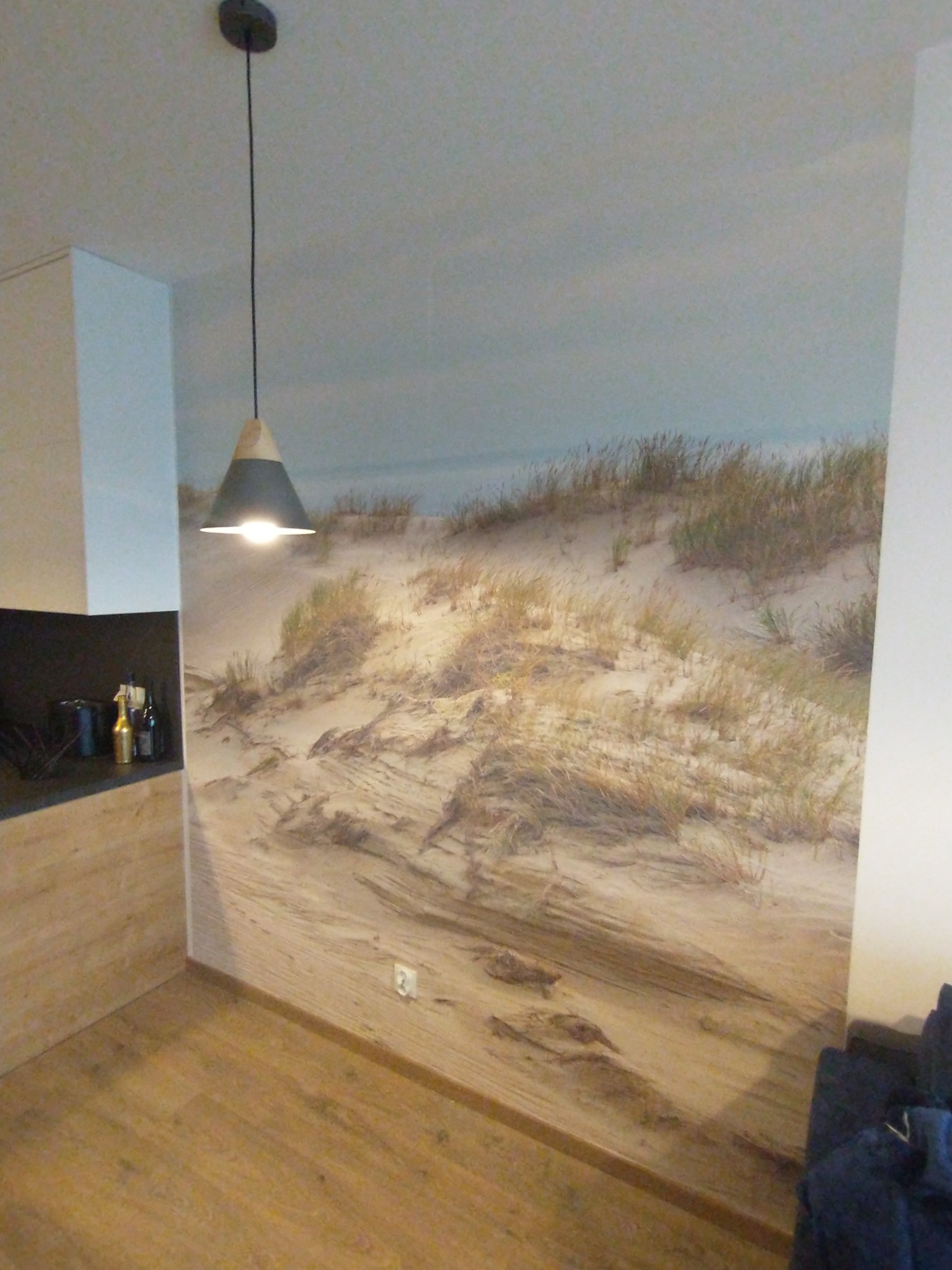 Wnętrze pokoju z fototapetą przedstawiającą wydmy z trawami, drewnianą podłogą, lampą wiszącą z abażurem w kształcie stożka i fragmentem zabudowy kuchennej.