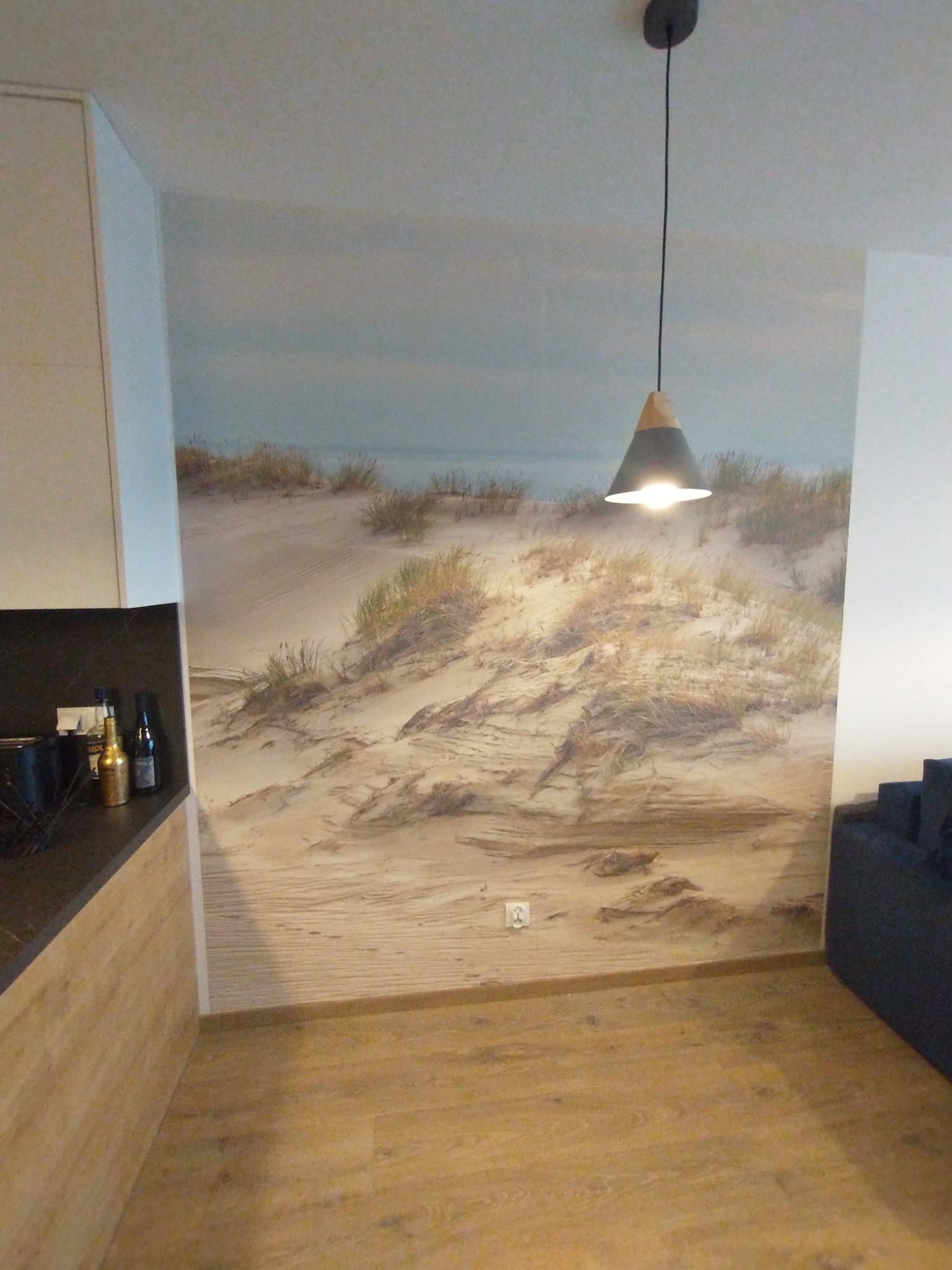 Wnętrze pokoju z fototapetą przedstawiającą piaszczyste wydmy, oświetlone nowoczesną lampą wiszącą i uzupełnione drewnianą podłogą oraz fragmentem mebli kuchennych.