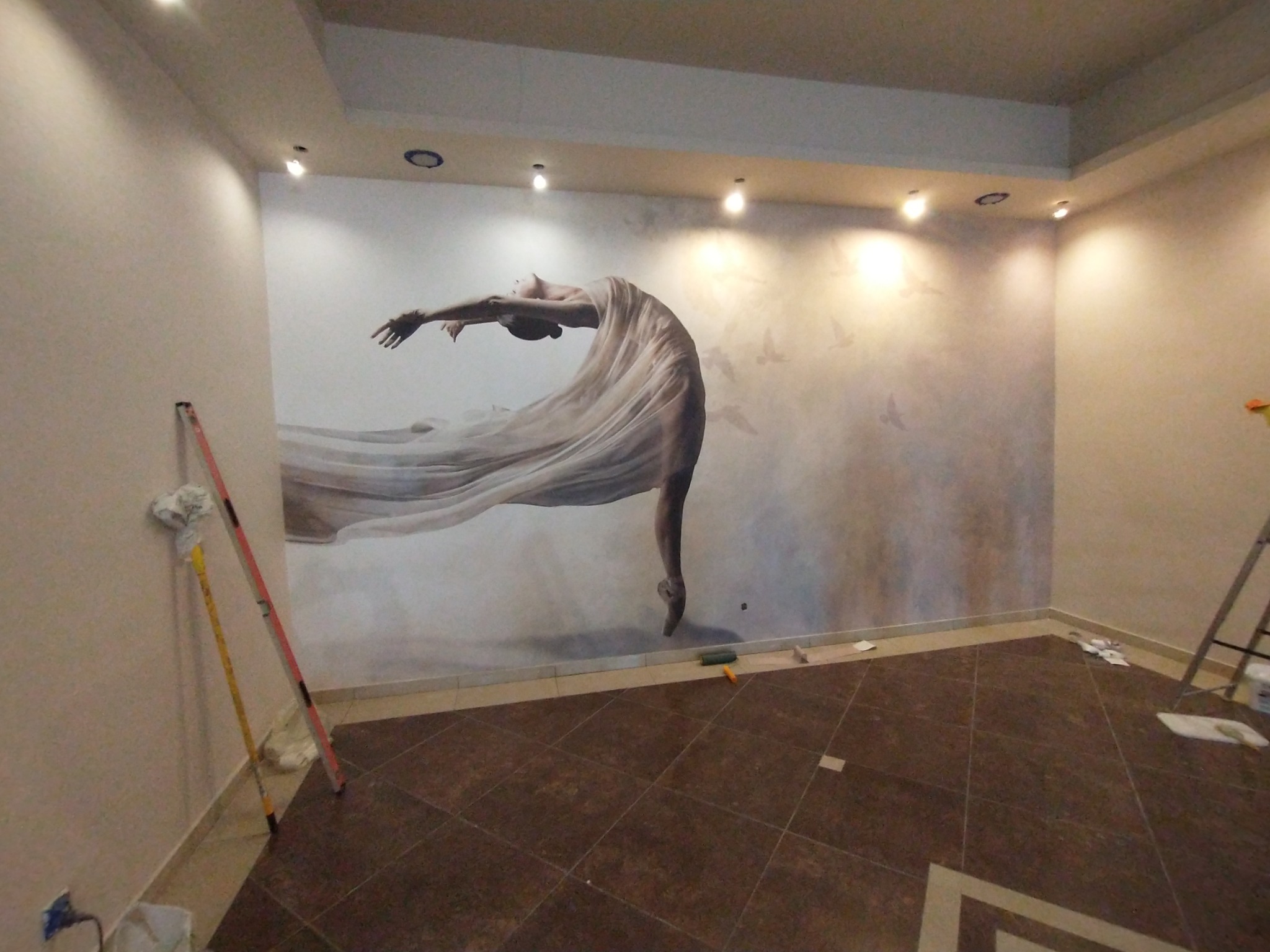 Ściana w pokoju z fototapetą przedstawiającą tancerkę w zwiewnej sukni, widoczne narzędzia malarskie i drabina, brązowa podłoga z płytek.