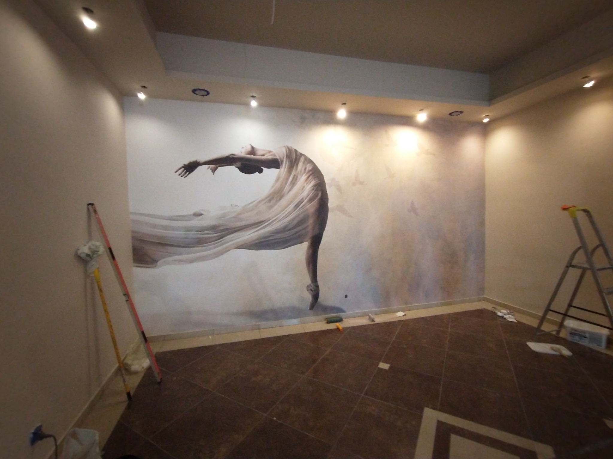 Wnętrze pokoju z fototapetą przedstawiającą tancerkę w zwiewnej sukni, widoczne narzędzia malarskie i drabina, podkreślające etap wykończenia.