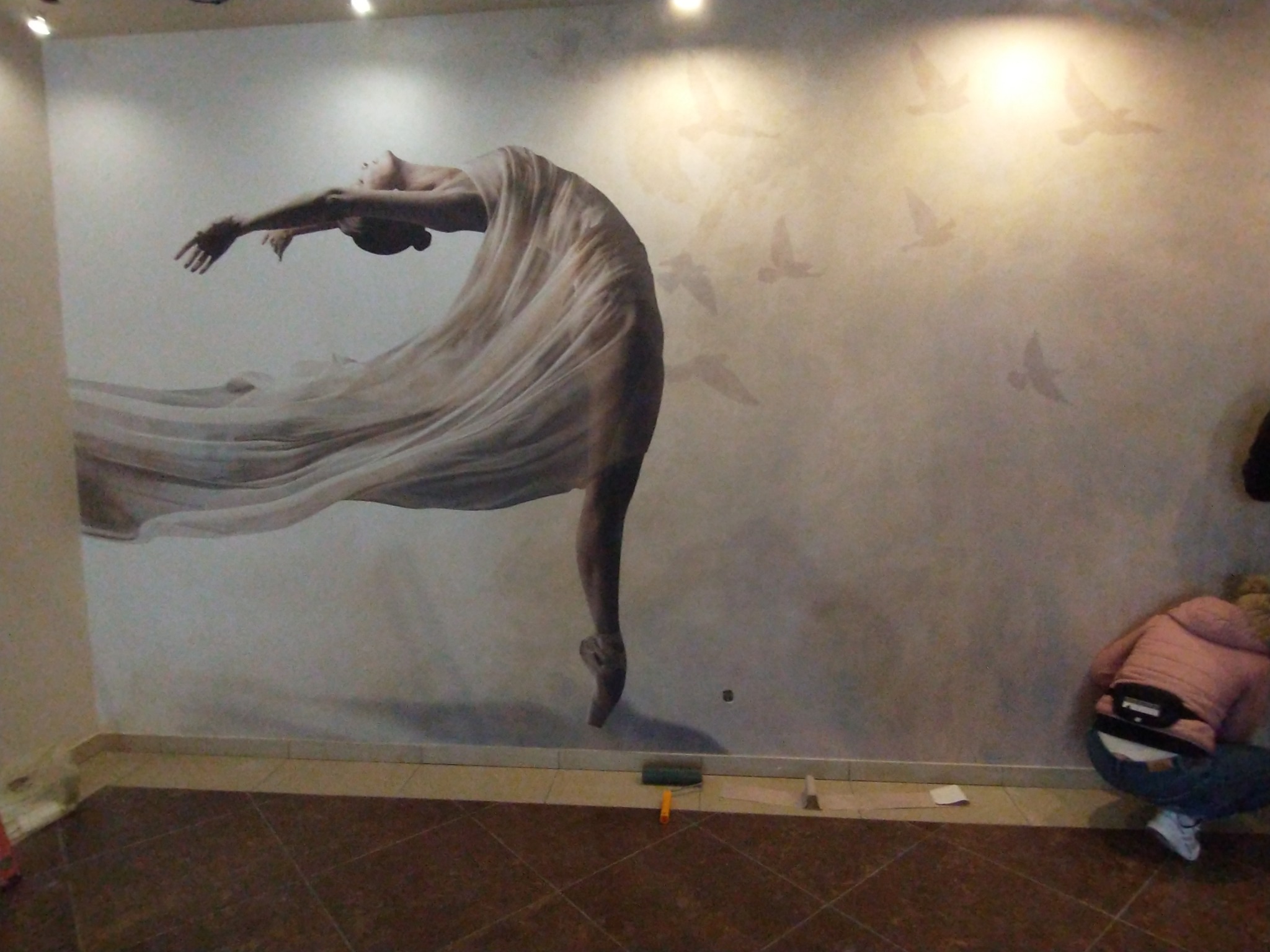 Naklejona na ścianie fototapeta z motywem baleriny w tańcu i ptaków w tle, widoczna osoba w trakcie pracy przy wygładzaniu tapety, na podłodze leżą narzędzia.