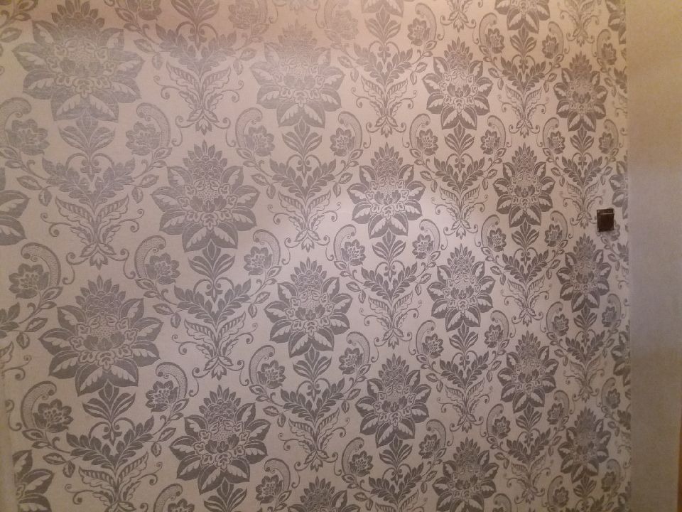 Ściana pokryta beżową tapetą w ornamenty roślinne w odcieniach szarości, z widocznym gniazdkiem elektrycznym.