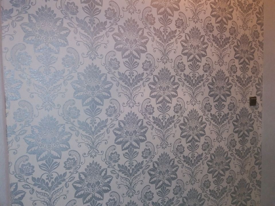 Ściana pokryta tapetą w kwiatowe, srebrzyste wzory na jasnym tle, widoczny fragment ściany z włącznikiem.