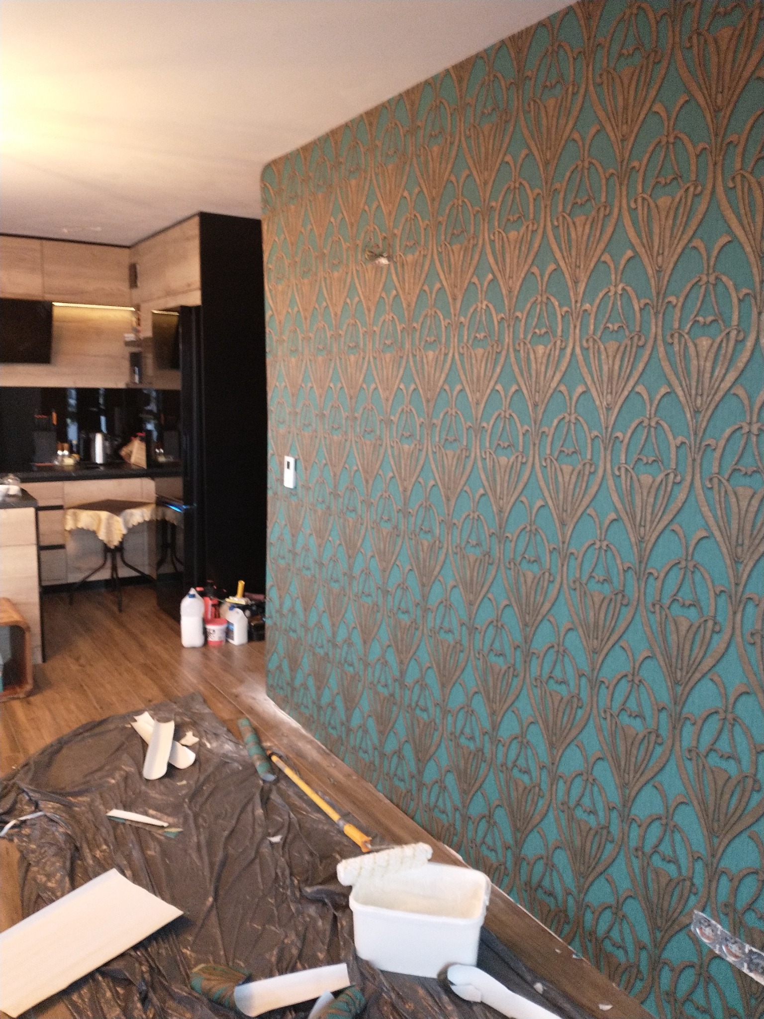 Ściana pokryta tapetą w stylu art deco, z turkusowym tłem i złotymi wzorami kwiatowymi, widoczne narzędzia i akcesoria do tapetowania na folii ochronnej na podłodze.