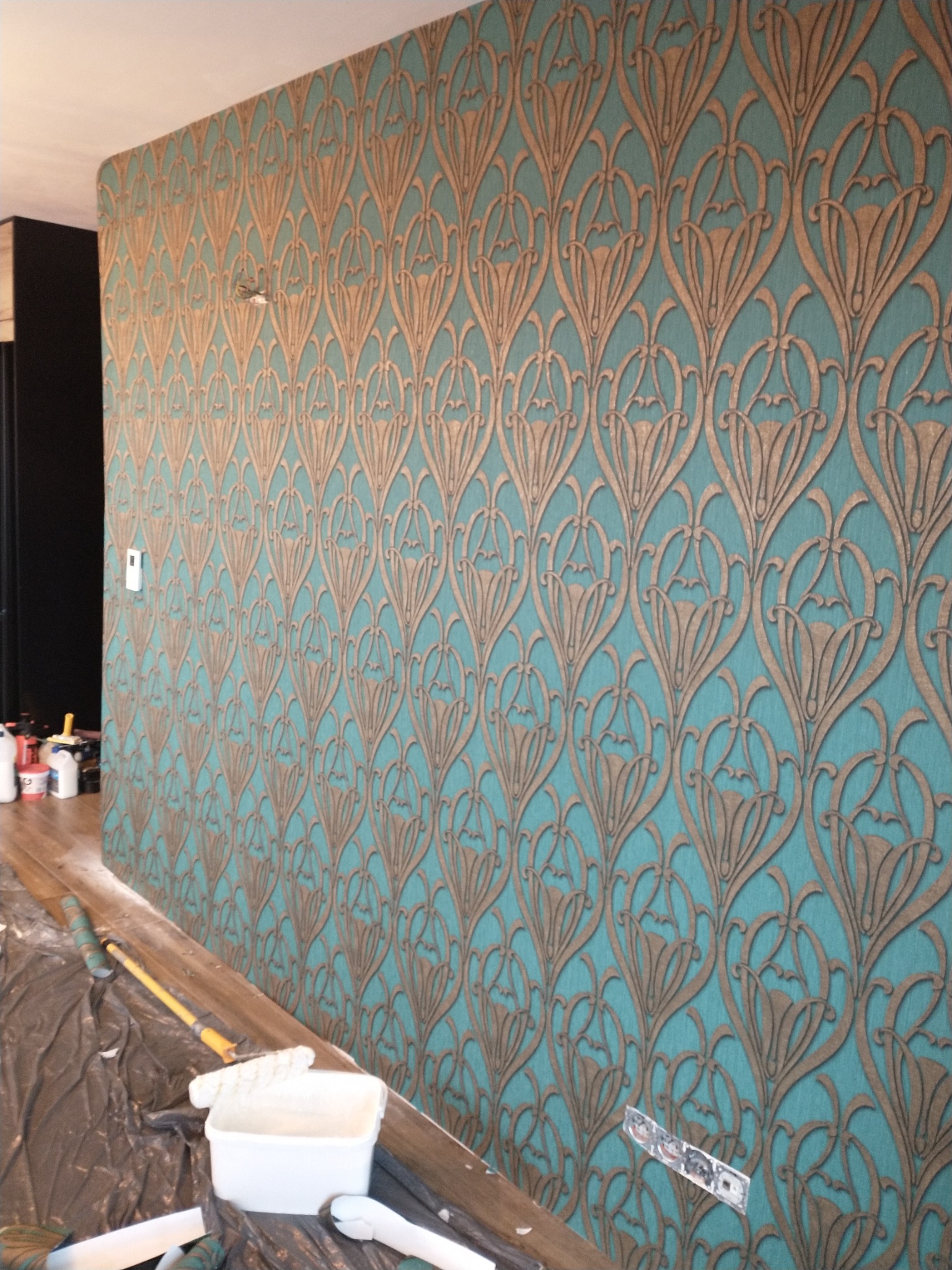 Ściana pokryta wzorzystą tapetą w odcieniach turkusu i złota, z widocznymi narzędziami i akcesoriami do tapetowania na pierwszym planie, w tym wiadro z klejem i wałek.