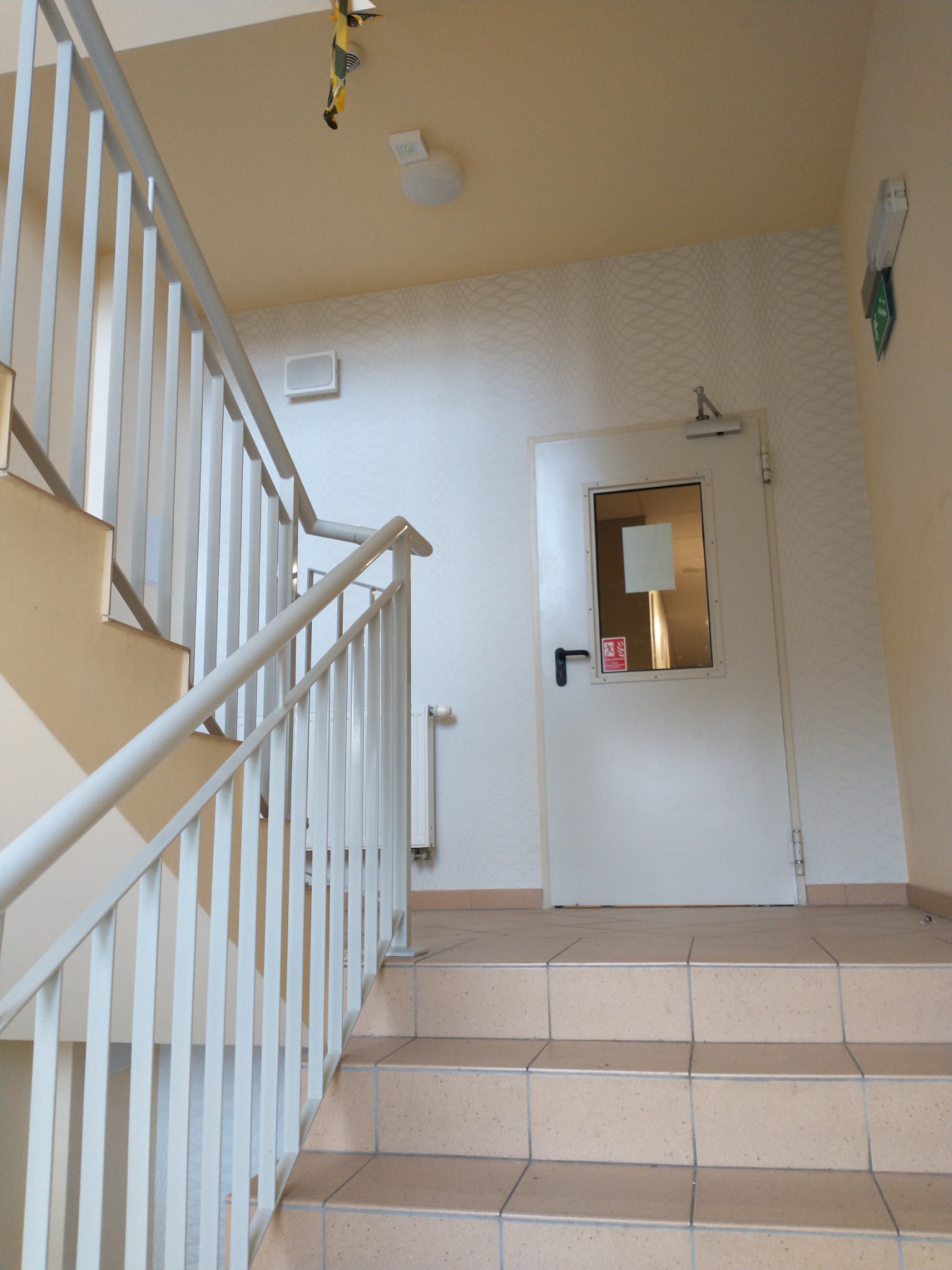 Klatka schodowa z jasnymi płytkami na schodach, białą balustradą i białą ścianą z delikatnym wzorem, prowadząca do białych drzwi z okienkiem.