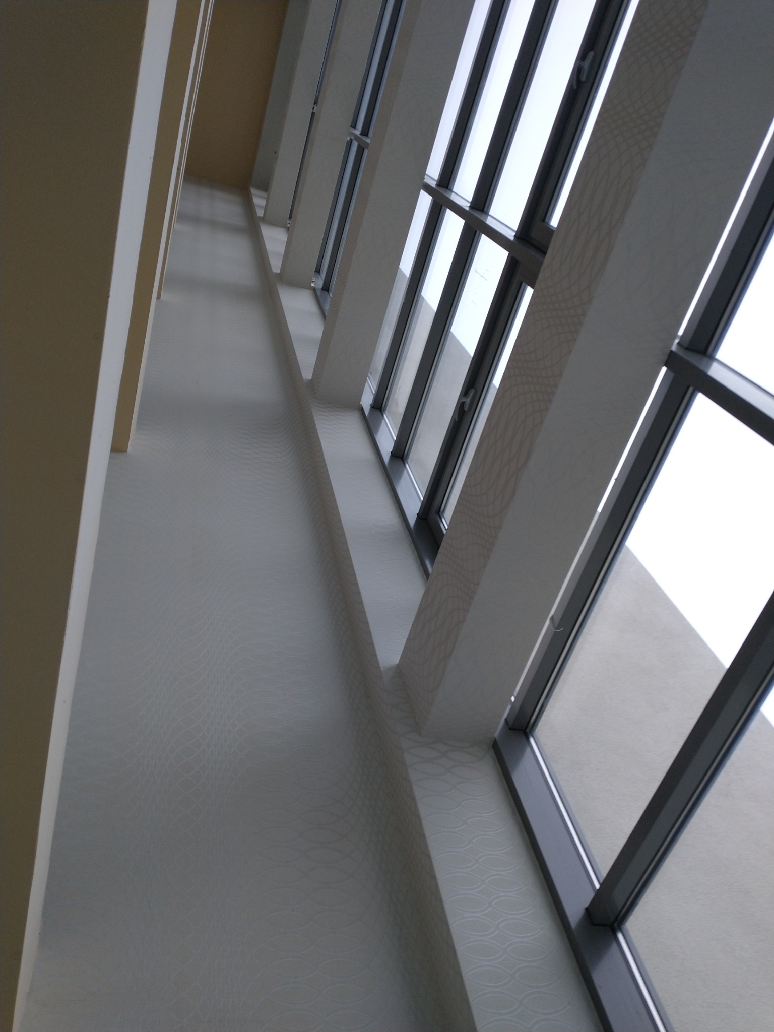 Wnętrze z wysokimi oknami i ścianą pokrytą tapetą o delikatnym wzorze w odcieniach bieli i szarości, widziane z dołu pod kątem.