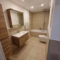 Nowoczesna łazienka z beżowymi płytkami, wiszącą szafką z umywalką z drewnianymi frontami, grzejnikiem drabinkowym i toaletą.