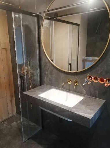 Nowoczesna łazienka w trakcie remontu: szara kamienna umywalka z złotą armaturą, okrągłe lustro w złotej ramie, widoczne elementy instalacji elektrycznej