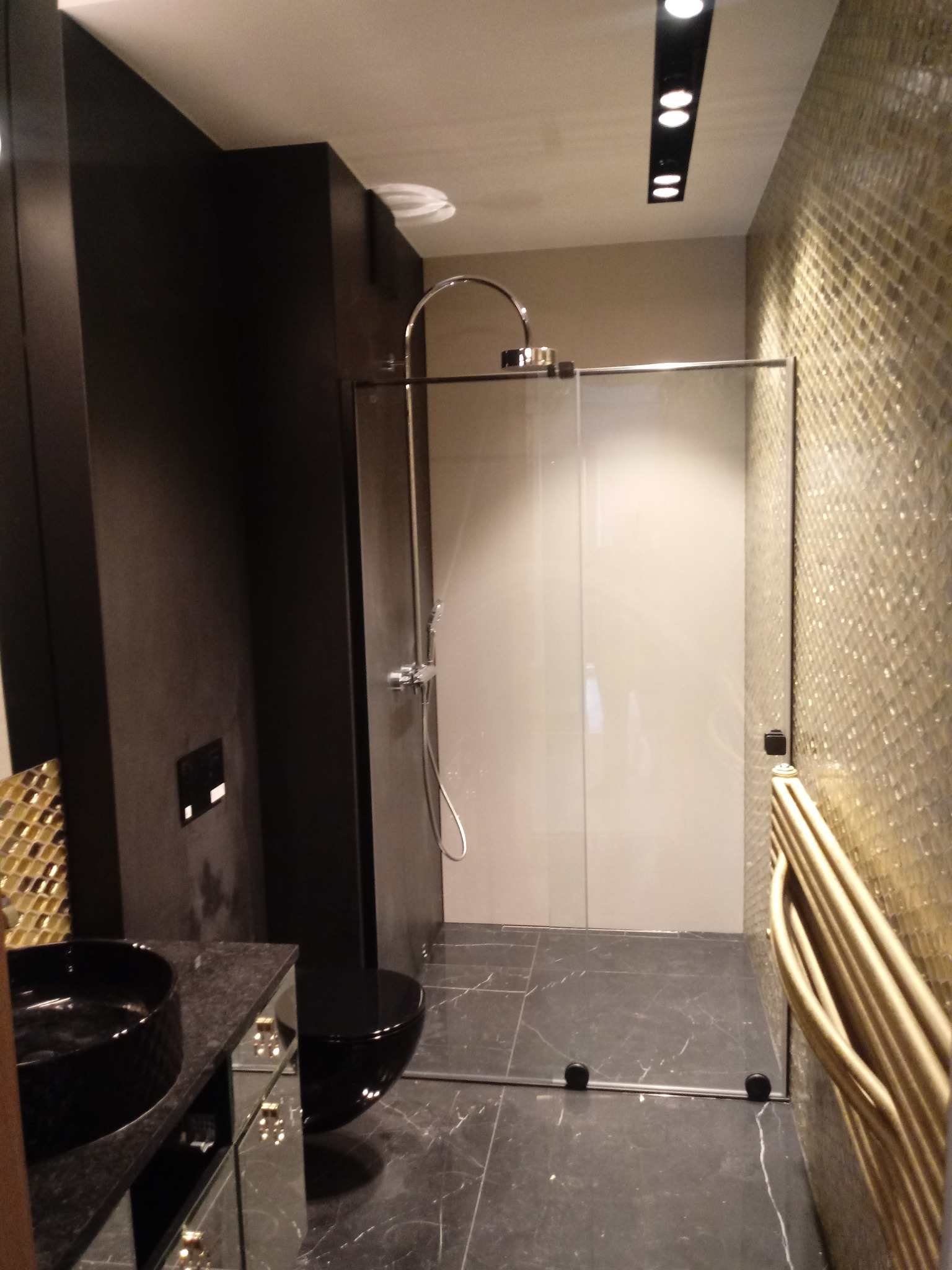 Nowoczesna łazienka z czarną umywalką nablatową, toaletą w zabudowie, kabiną prysznicową ze szklanymi drzwiami przesuwnymi i złotą mozaiką na ścianie.