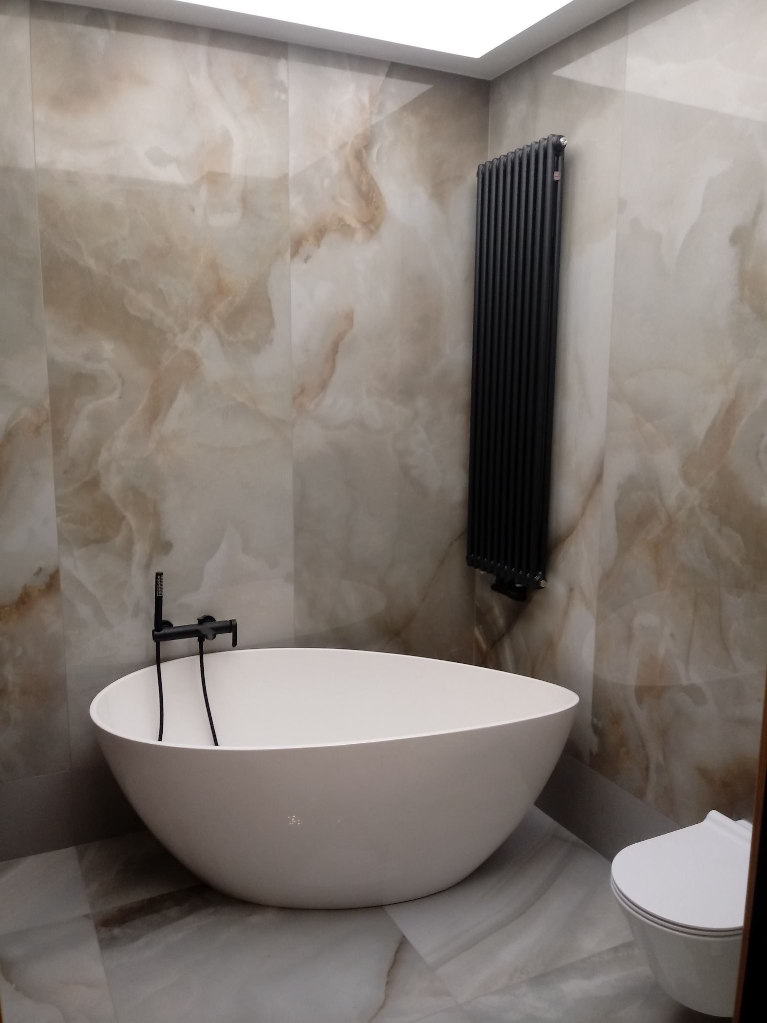Nowoczesna łazienka z wolnostojącą białą wanną, czarną armaturą i grzejnikiem drabinkowym na tle marmurowych płytek.