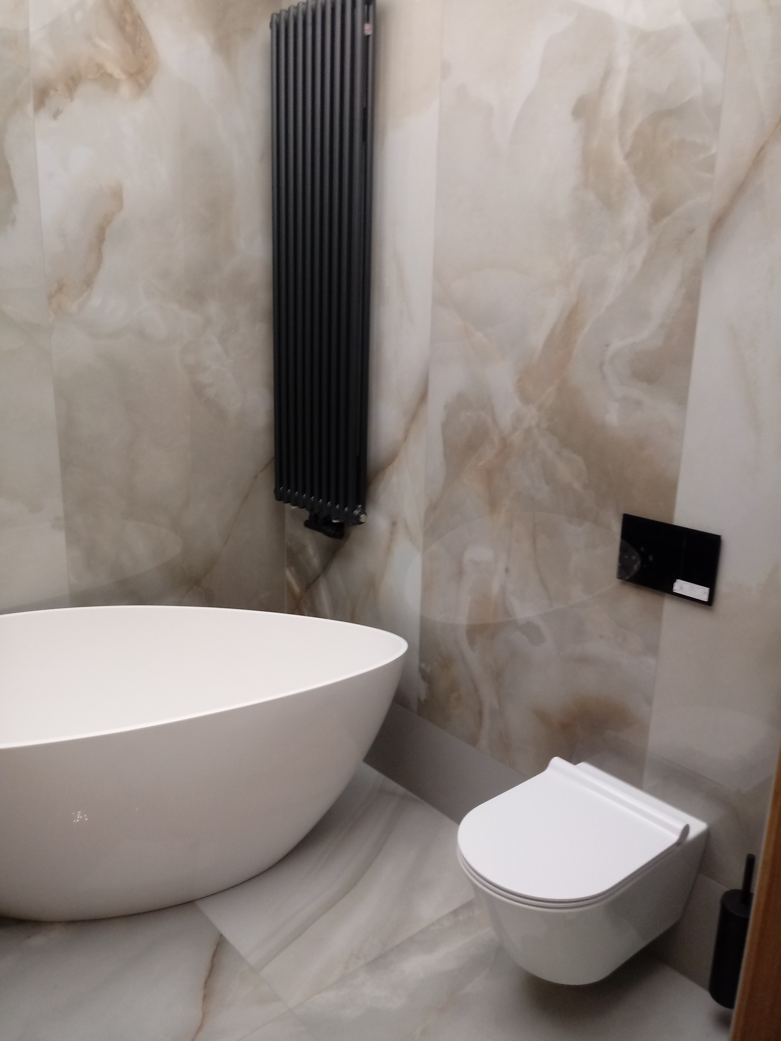 Nowoczesna łazienka z owalną wanną, podwieszaną toaletą i pionowym, czarnym grzejnikiem na tle marmurowych płytek.