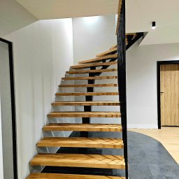 POLMAKS Schody Szyte Ogniem - Spiralne schody z drewnianymi stopniami i czarną metalową balustradą w nowoczesnym wnętrzu, widok z dołu, z widocznym fragmentem podłogi z szarych płytek i drewnianej podłogi w tle.