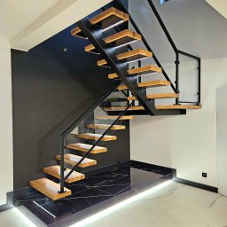 POLMAKS Schody Szyte Ogniem - Nowoczesne schody z drewnianymi stopniami, metalową konstrukcją i szklanymi balustradami. Podświetlenie LED dodaje elegancji. Czarna ściana i marmurowa podłoga.