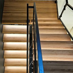 POLMAKS Schody Szyte Ogniem - Nowoczesne schody z drewna z podświetleniem LED, czarną metalową balustradą i szklanymi panelami, widok z góry. Detal wykończenia wnętrza.