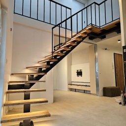 POLMAKS Schody Szyte Ogniem - Nowoczesne schody z drewnianymi stopniami i czarną metalową konstrukcją, podświetlane LED, w jasnym wnętrzu z antresolą i oknem dachowym.