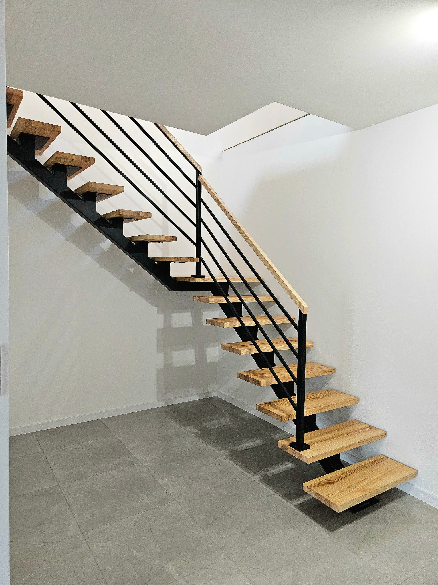 Nowoczesne schody z drewnianymi stopniami i czarną metalową konstrukcją, minimalistyczne wnętrze z szarą podłogą z płytek.