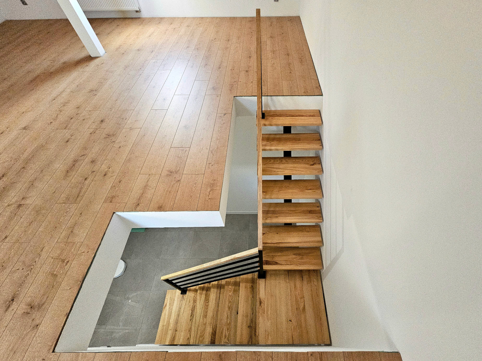 Drewniane schody z czarnymi metalowymi elementami, widok z góry, z drewnianą podłogą na górnym poziomie i szarymi płytkami na dolnym poziomie.