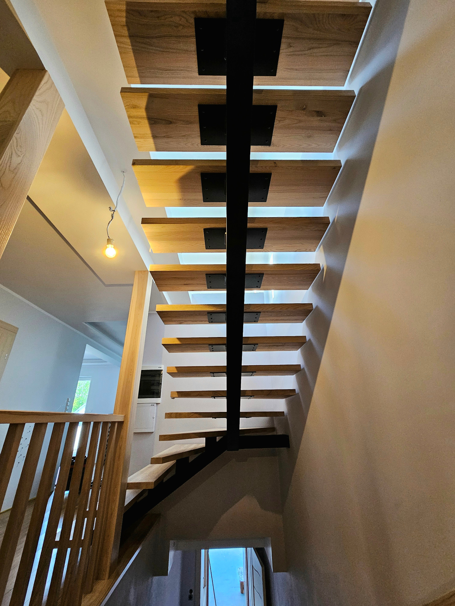 Nowoczesne schody z drewnianymi stopniami mocowanymi do centralnej metalowej belki, widok od dołu, z widocznym oświetleniem pod stopniami i drewnianą balustradą.