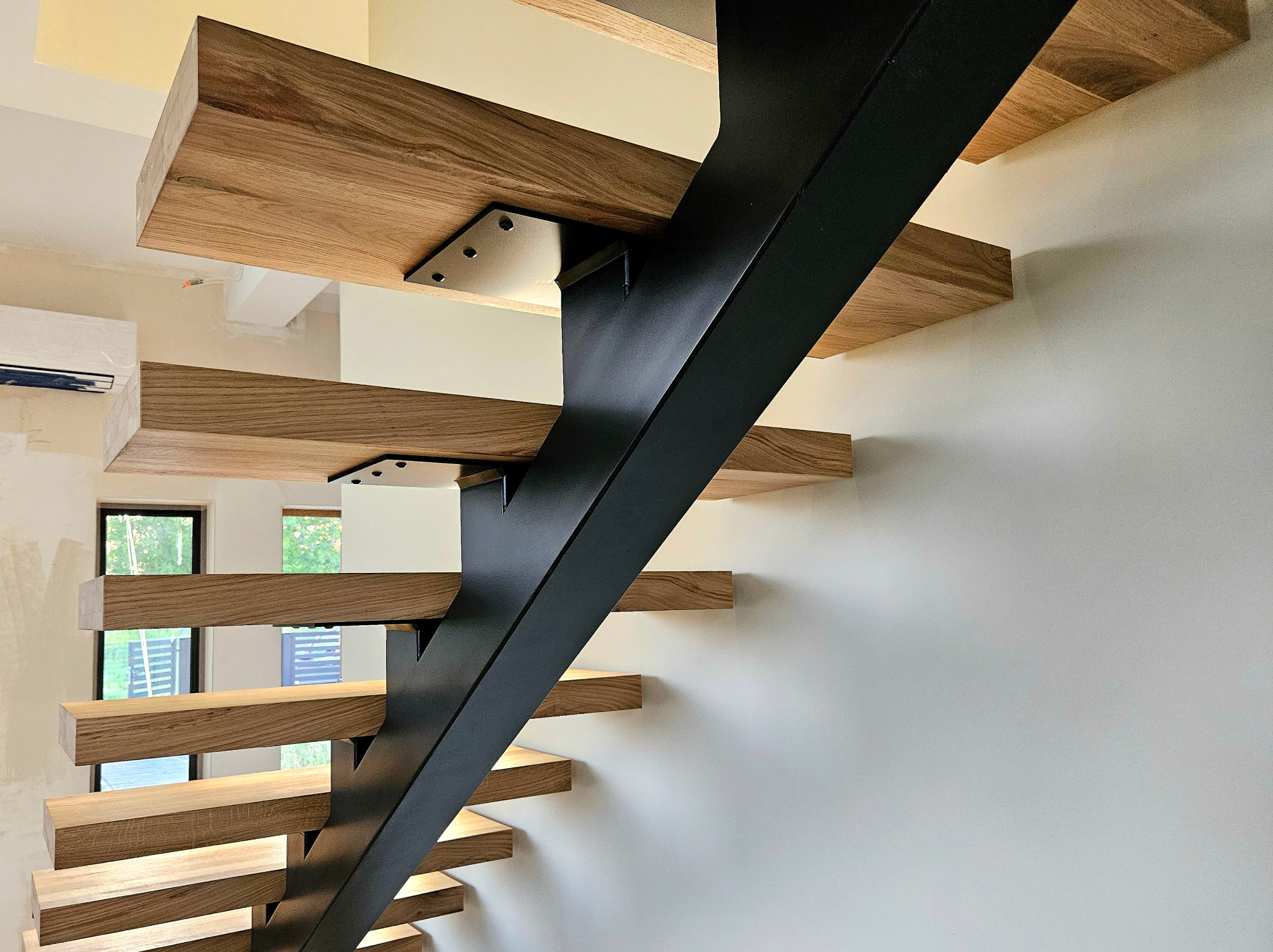 Nowoczesne schody wspornikowe z drewnianymi stopniami i czarną, ukośną konstrukcją nośną przy białej ścianie, widoczne okno w tle.