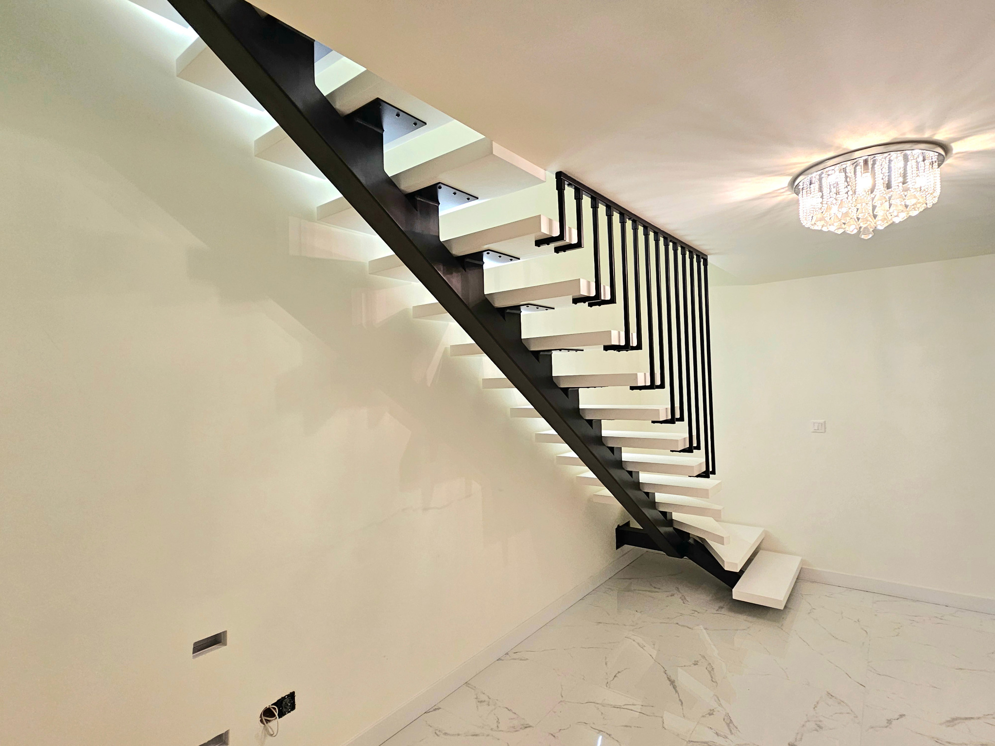 Nowoczesne schody z białymi stopniami i czarną metalową konstrukcją, oświetlone od spodu, w minimalistycznym wnętrzu z marmurową podłogą i kryształowym żyrandolem.