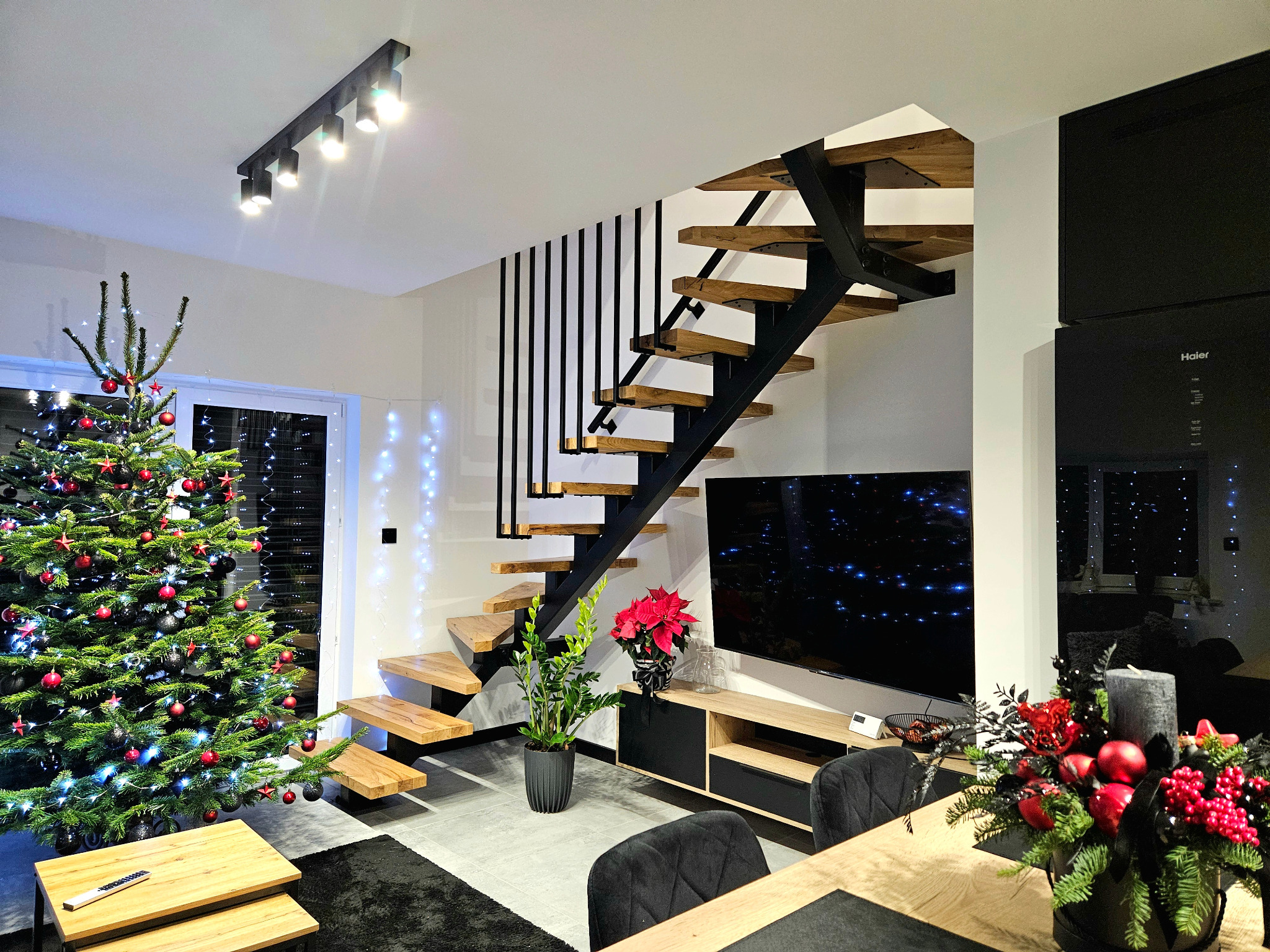Nowoczesne schody z drewnianymi stopniami i czarną metalową konstrukcją w jasnym wnętrzu, z choinką w tle.