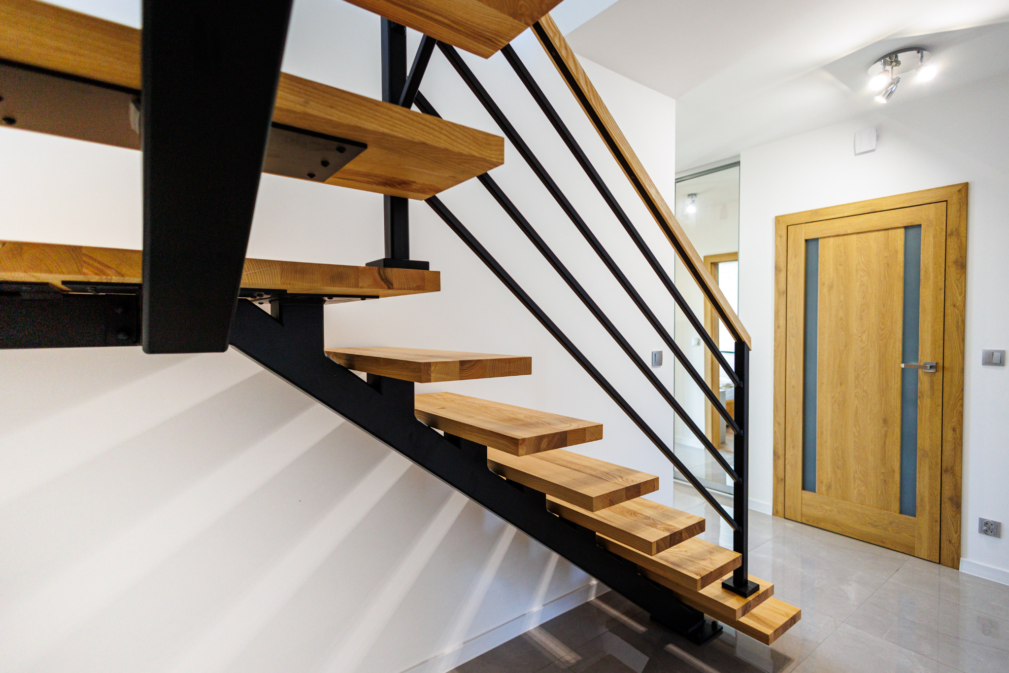 Nowoczesne schody z drewnianymi stopniami i czarną metalową konstrukcją, widoczne z dołu, z drewnianymi drzwiami w tle.