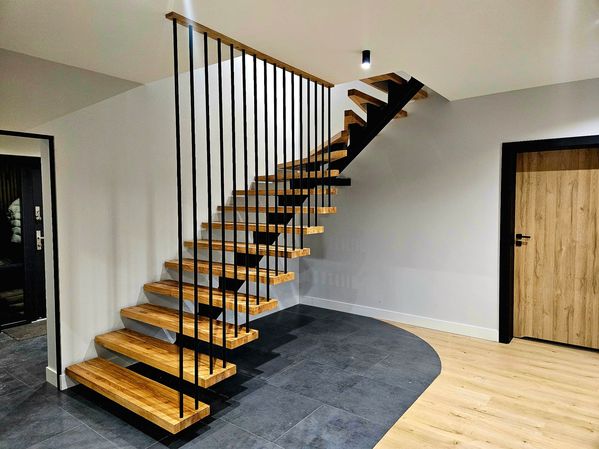 Nowoczesne schody wspornikowe z drewnianymi stopniami i czarnymi, pionowymi balustradami, w minimalistycznym wnętrzu z szarą ścianą i kontrastującą podłogą z płytek w dwóch odcieniach.