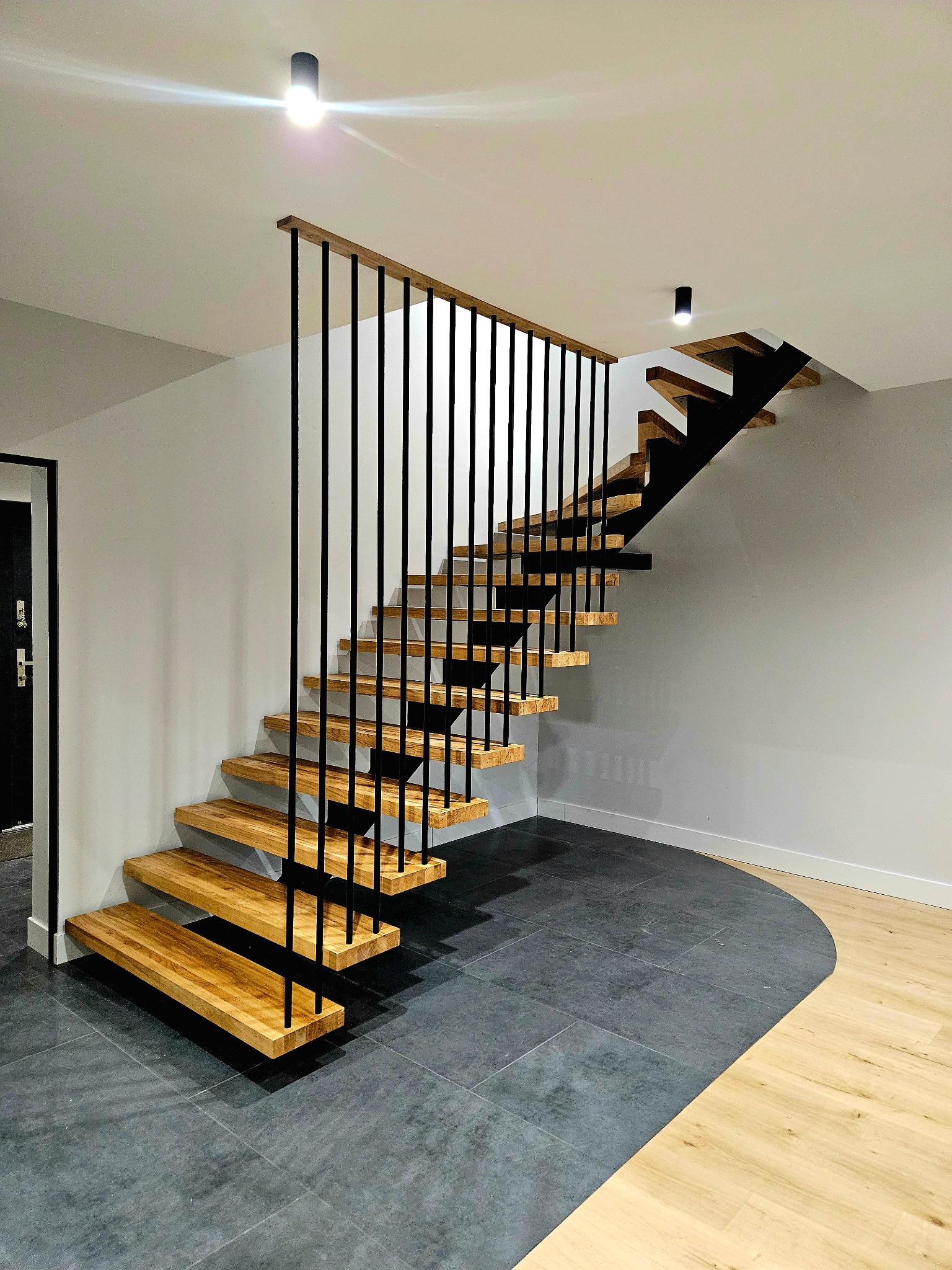 Nowoczesne schody metalowe z drewnianymi stopniami i czarnymi, pionowymi balustradami w domu w Kleosinie, widok z dołu na tle szarej ściany i kontrastującej podłogi z płytek i paneli.