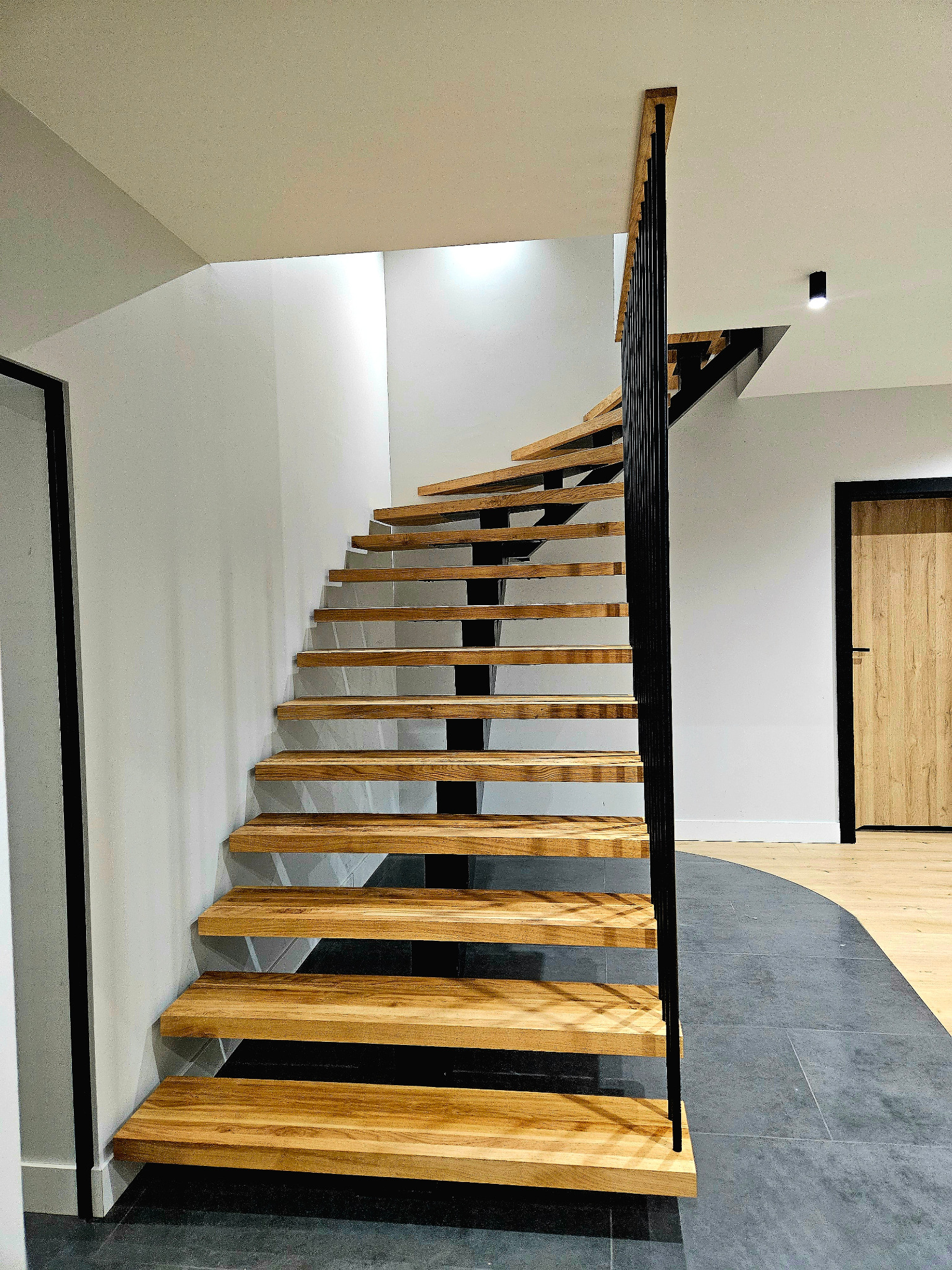 Spiralne schody z drewnianymi stopniami i czarną metalową balustradą w nowoczesnym wnętrzu, widok z dołu, z widocznym fragmentem podłogi z szarych płytek i drewnianej podłogi w tle.