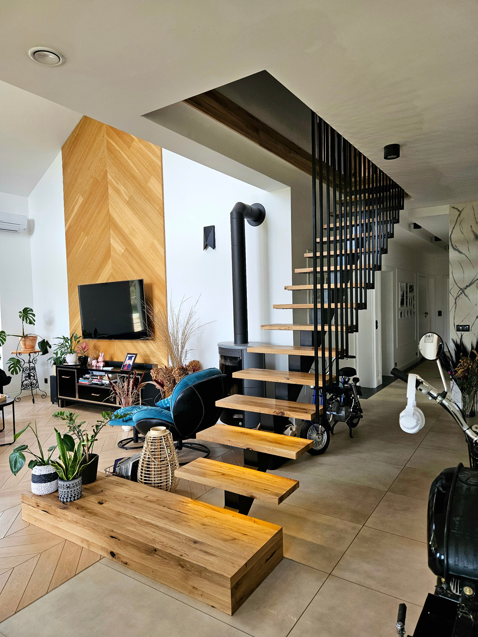 Nowoczesne schody z drewnianymi stopniami i czarną metalową balustradą w jasnym wnętrzu z elementami drewna i motocyklem w tle.