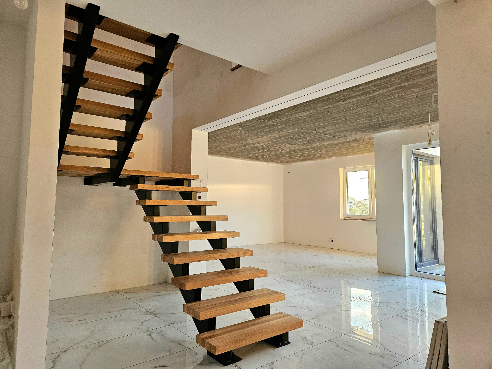 Nowoczesne schody z drewnianymi stopniami i czarną metalową konstrukcją w jasnym wnętrzu z marmurową podłogą i surowym betonowym sufitem.