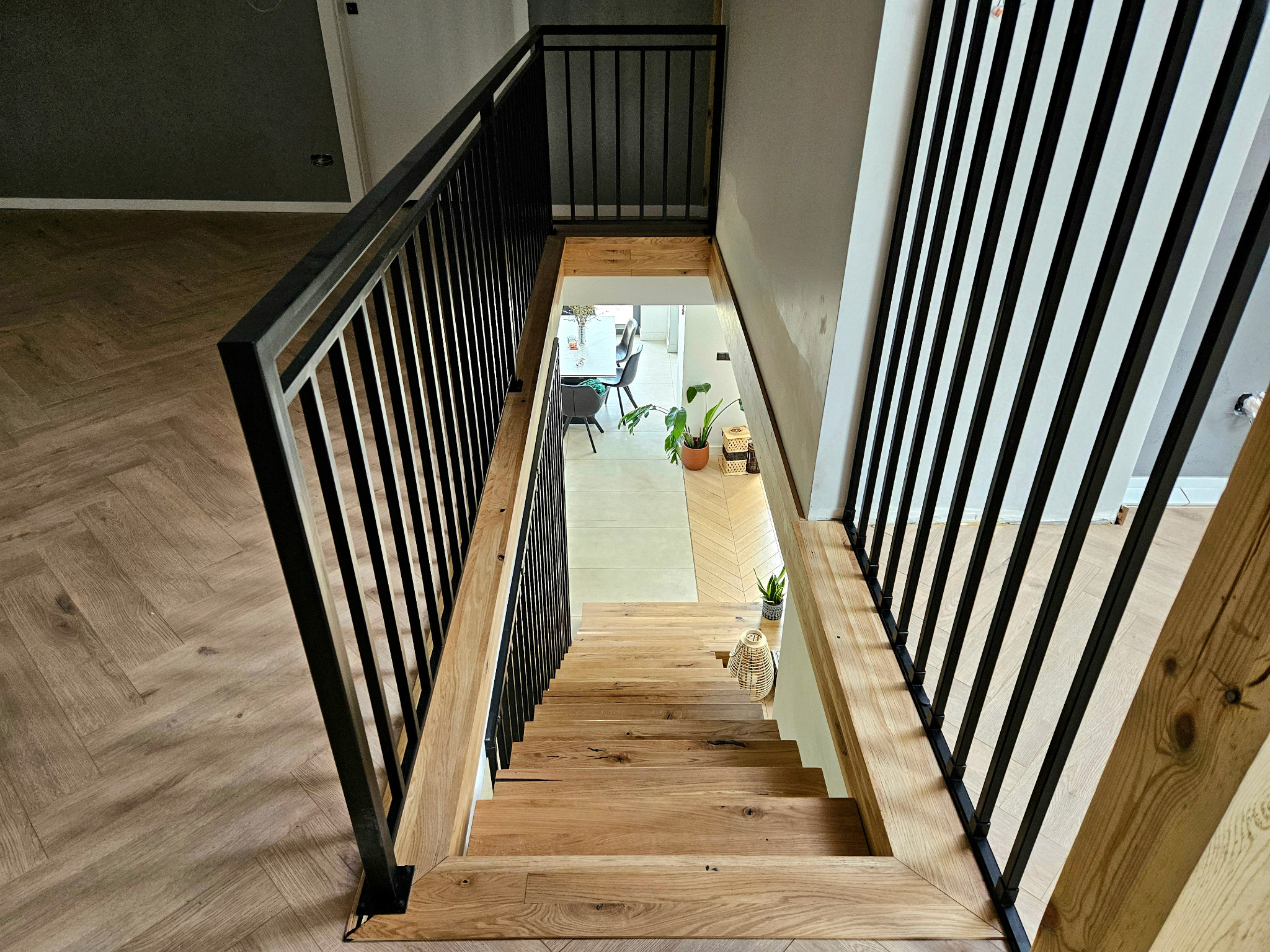 Nowoczesne schody z drewnianymi stopniami i czarną, metalową balustradą. Widok z góry na schody w jasnym wnętrzu z parkietem w jodełkę.