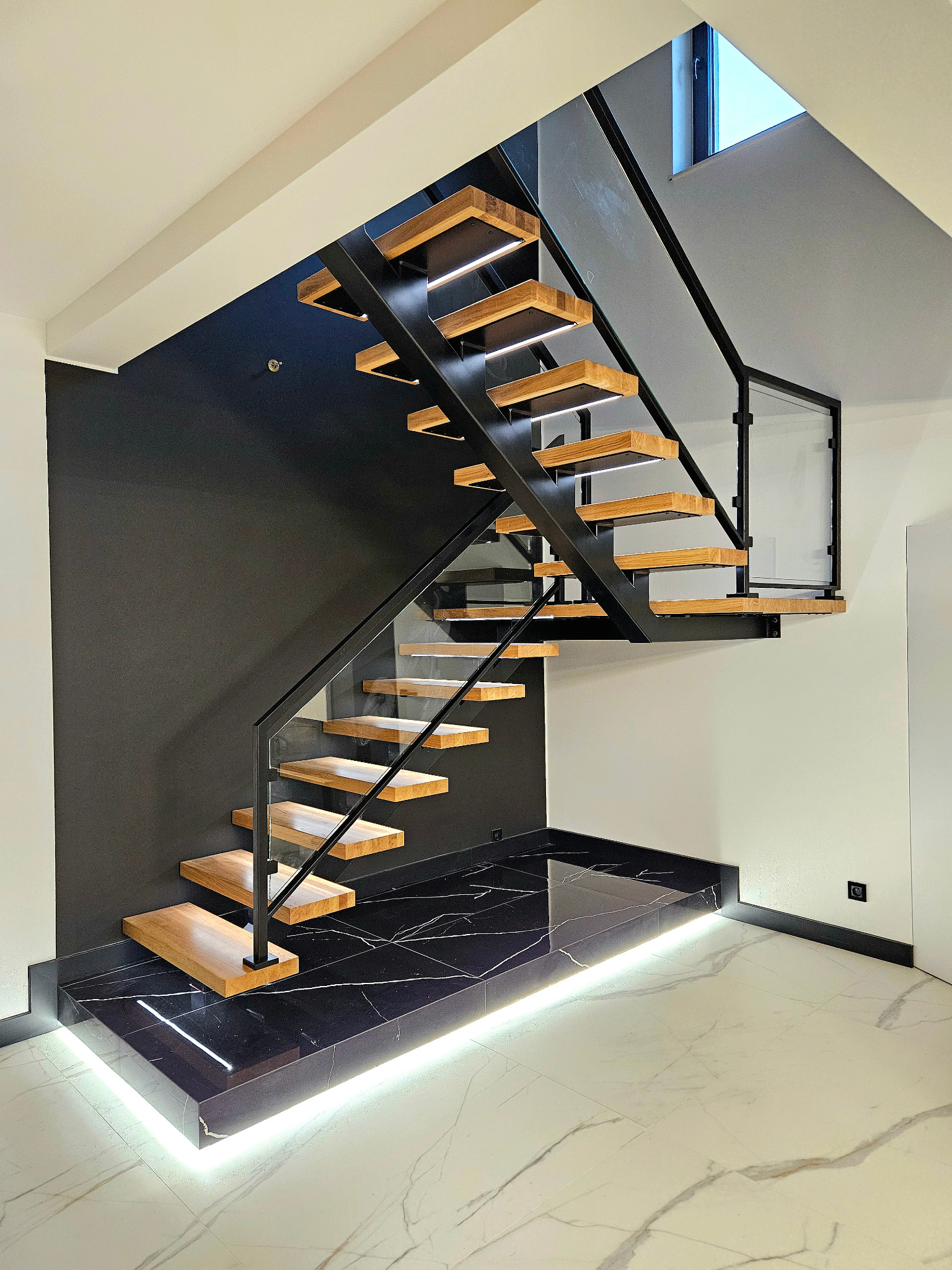 Nowoczesne schody z drewnianymi stopniami, metalową konstrukcją i szklanymi balustradami. Podświetlenie LED dodaje elegancji. Czarna ściana i marmurowa podłoga.