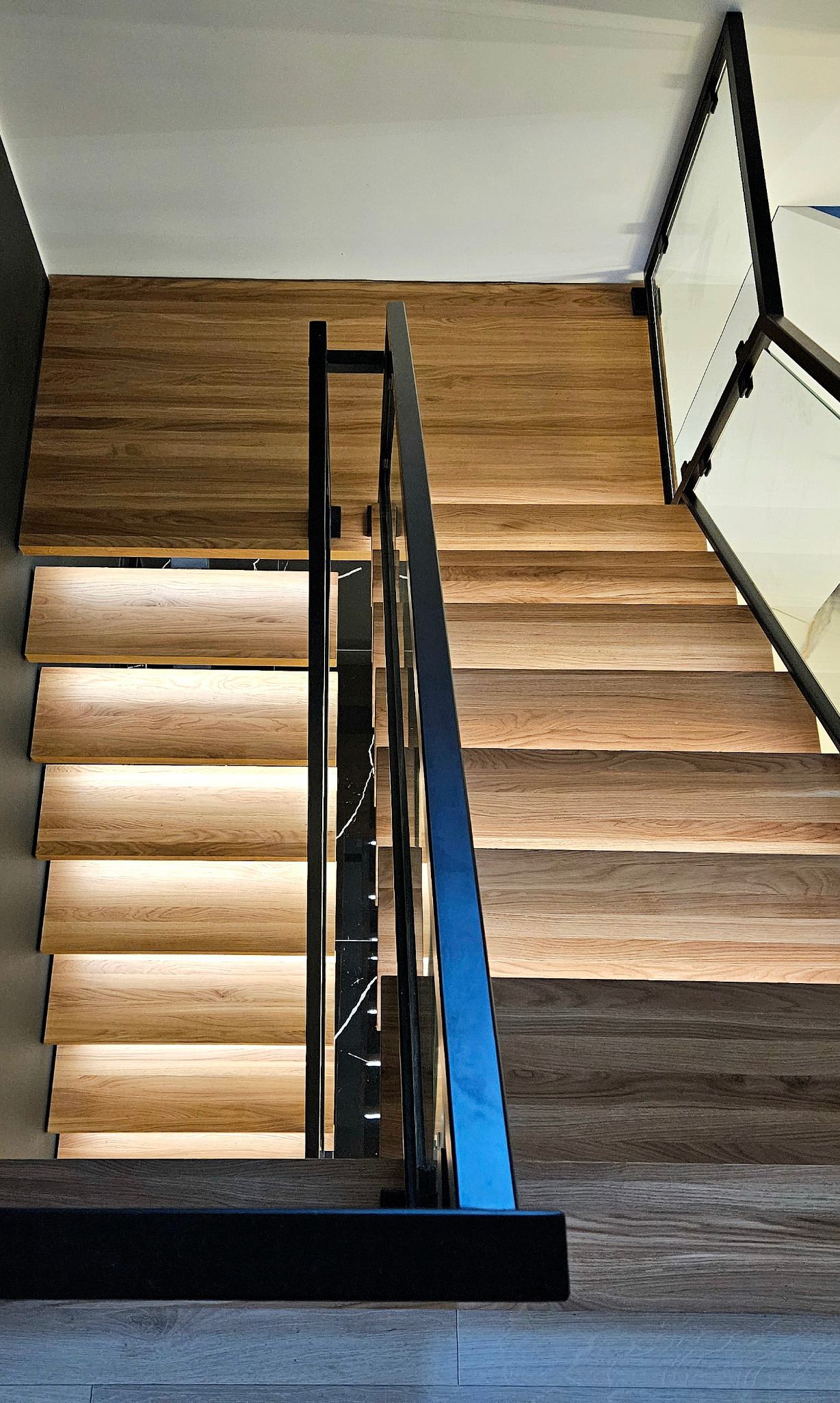 Nowoczesne schody z drewna z podświetleniem LED, czarną metalową balustradą i szklanymi panelami, widok z góry. Detal wykończenia wnętrza.