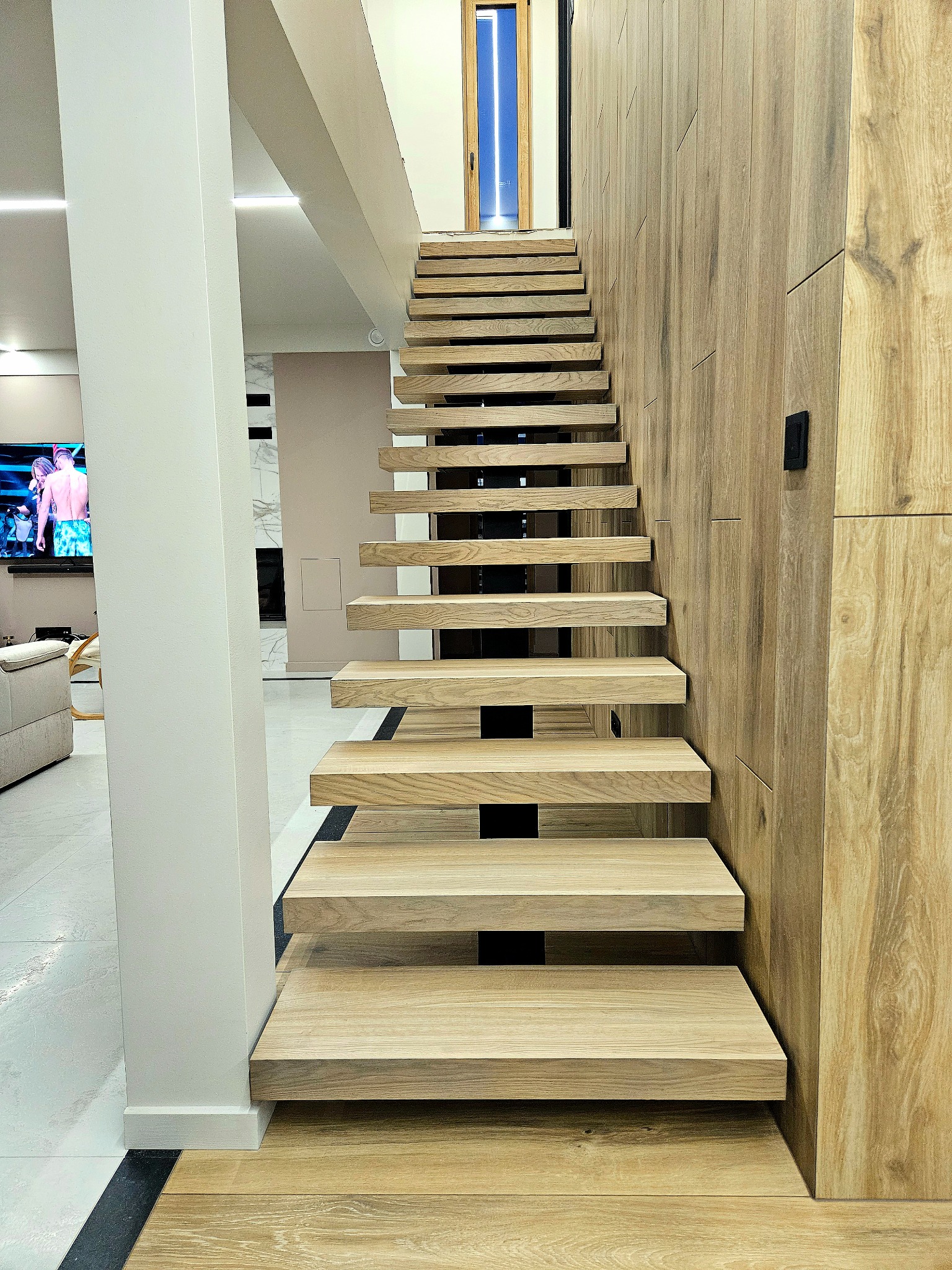 Drewniane schody wspornikowe z czarnymi elementami w nowoczesnym wnętrzu domu w Kleosinie. Ściana wykończona panelami drewnopodobnymi.